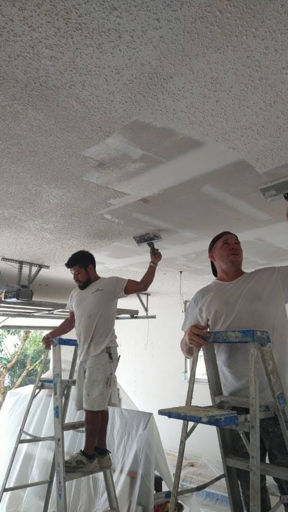 Painters on ladders scraping acoustic popcorn ceiling texture with broad knives in Naples home