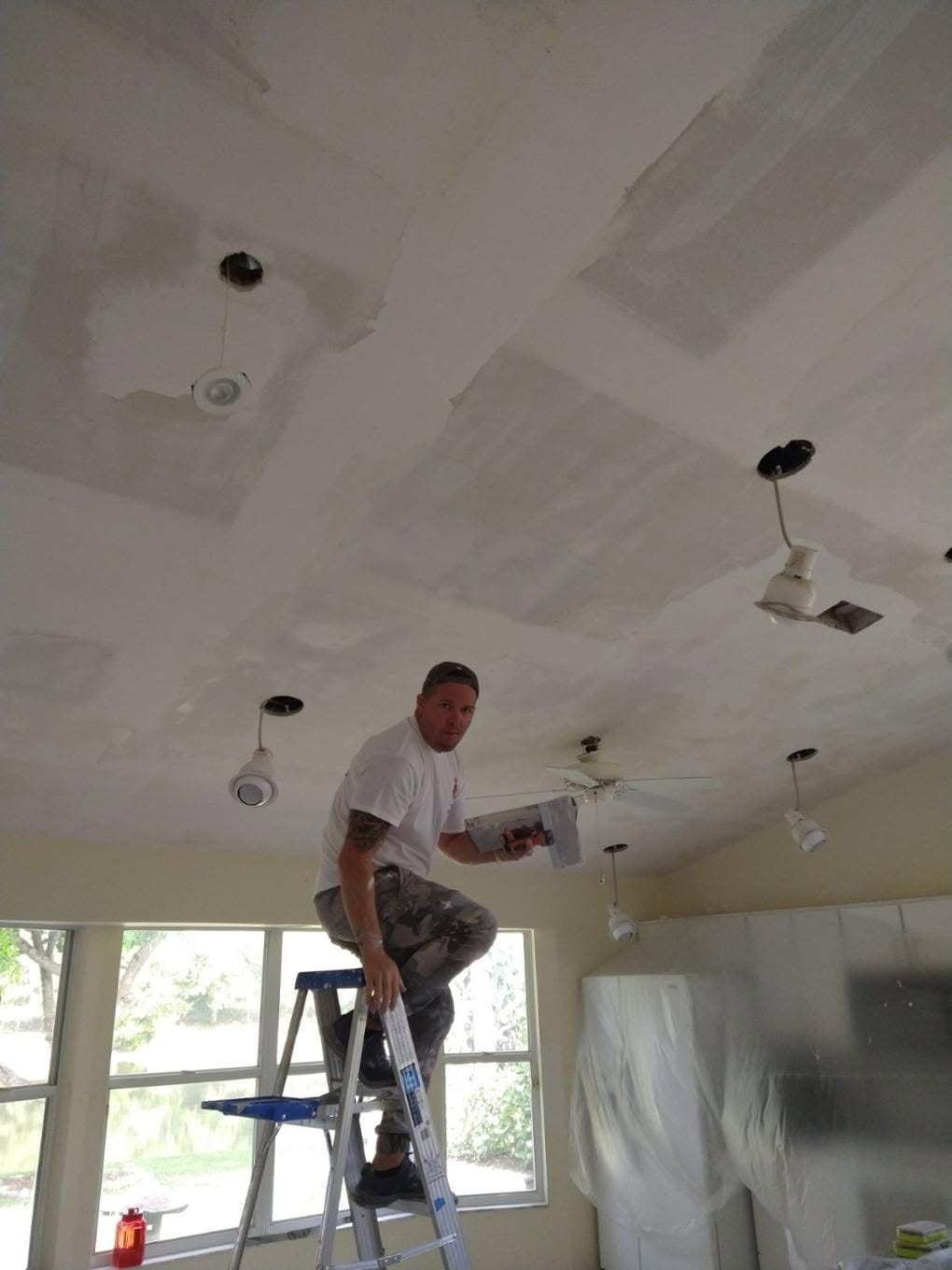 Painter holding drywall sander inspecting skimmed and smoothed ceiling around light fixtures in Naples living room