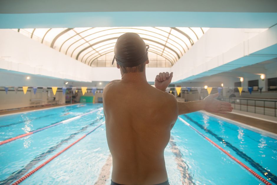 Swimmer in a cap stretching beside an indoor lap pool with lane lines and pennant flags