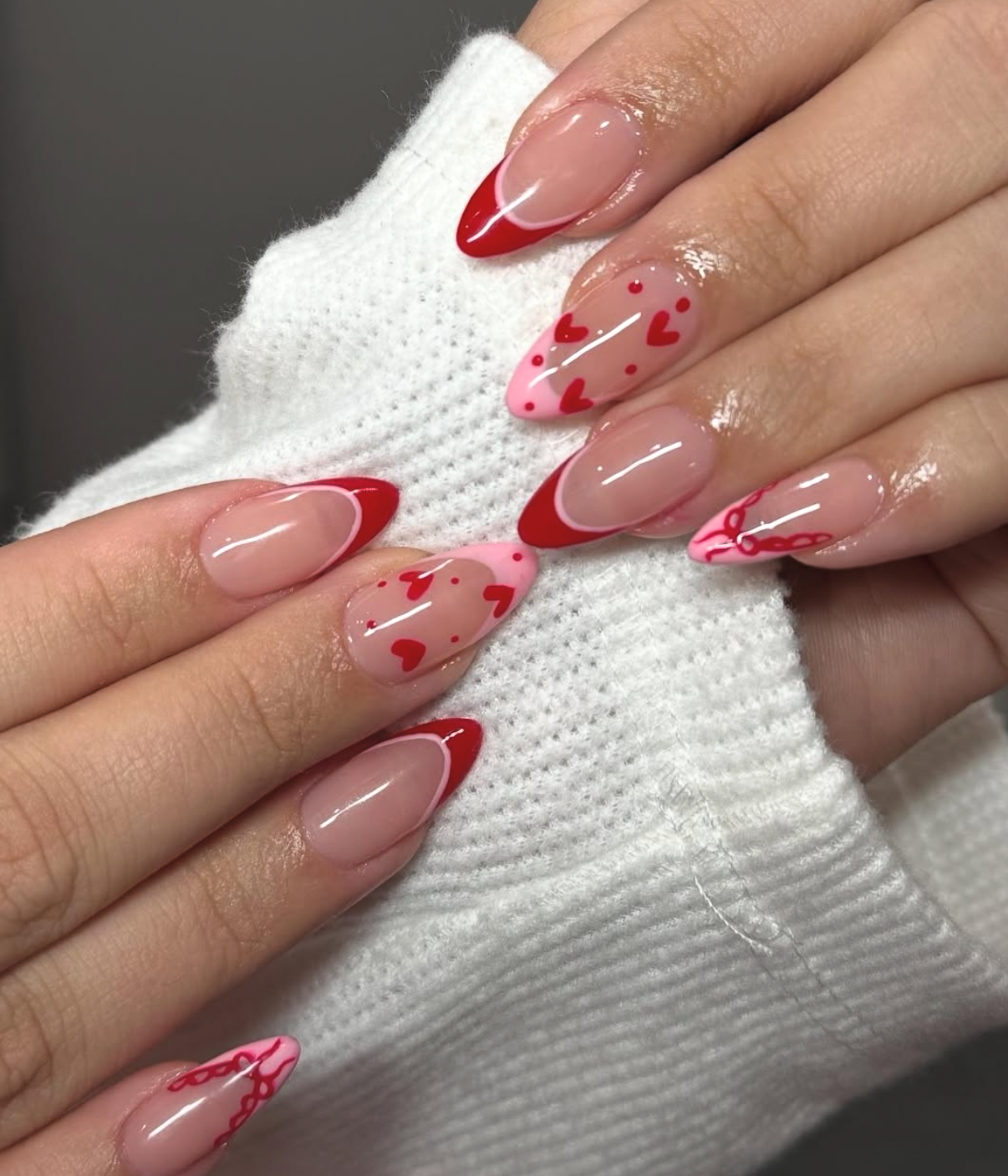 Almond nails with glossy red french tips, delicate heart motifs, and chain link ribbon details