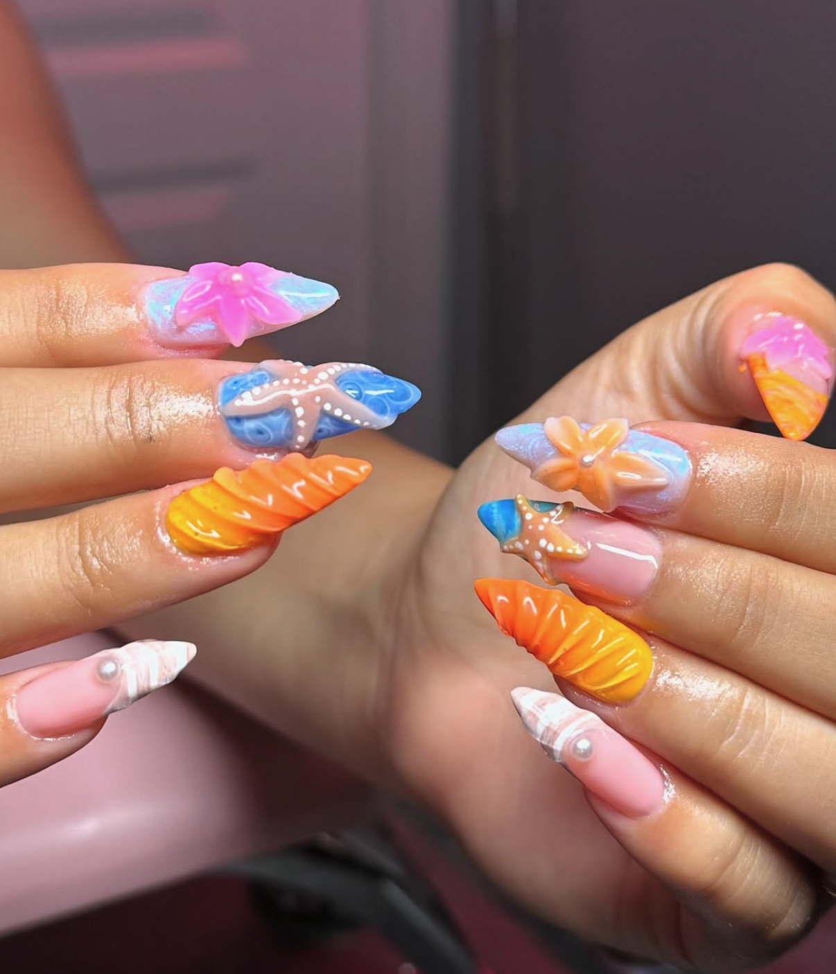Sculpted 3D beach nails with vibrant orange ribbed seashells, blue ocean water with white starfish and pearls, and pink tropical hibiscus
