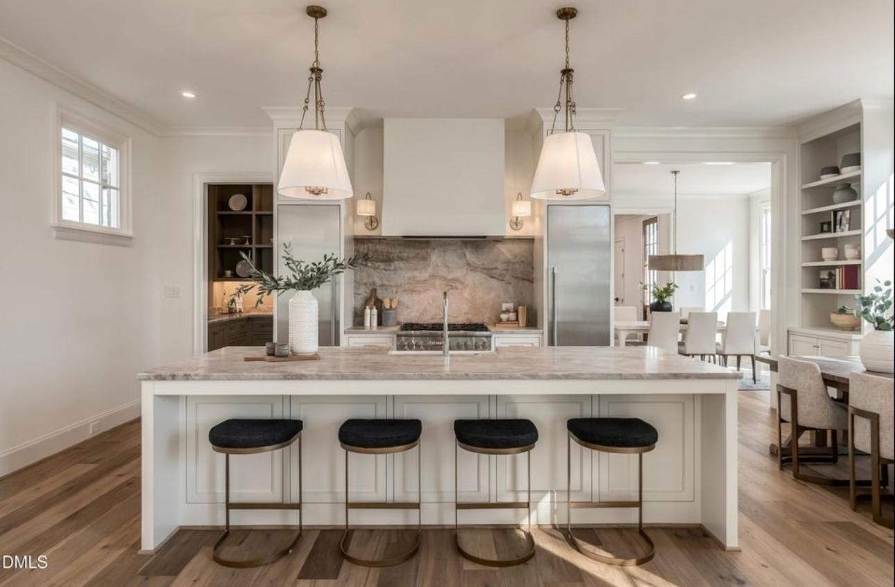 Custom luxury kitchen featuring a large marble-topped island with brass-accented counter stools, twin linen pendant lanterns, custom range hood, and open-plan dining space