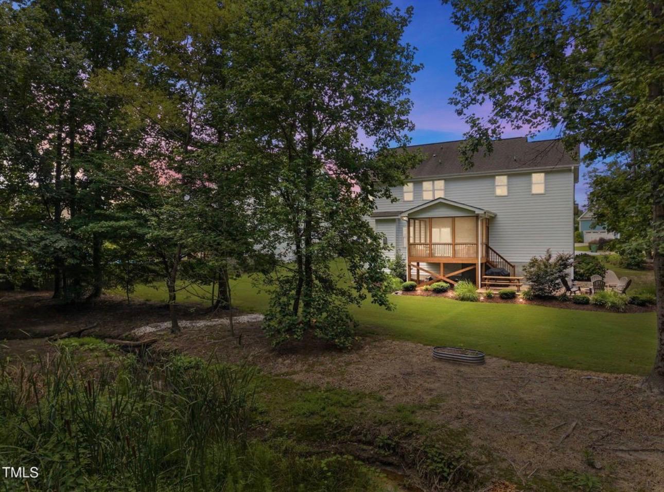 Twilight view of a Raleigh home with an illuminated elevated screened back porch overlooking a private lawn, woodland buffer, and twilight sky — represented and sold by Leah Beeson Real Estate