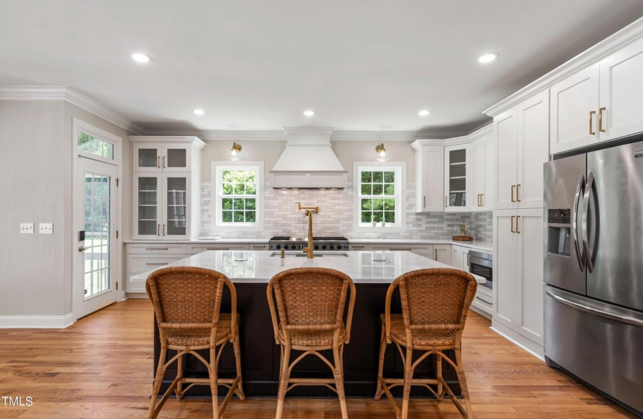 Bright open-concept kitchen with contrasting espresso island, white quartz counters, woven wicker barstools, brass sconces, and custom range hood — represented and sold by Leah Beeson Real Estate