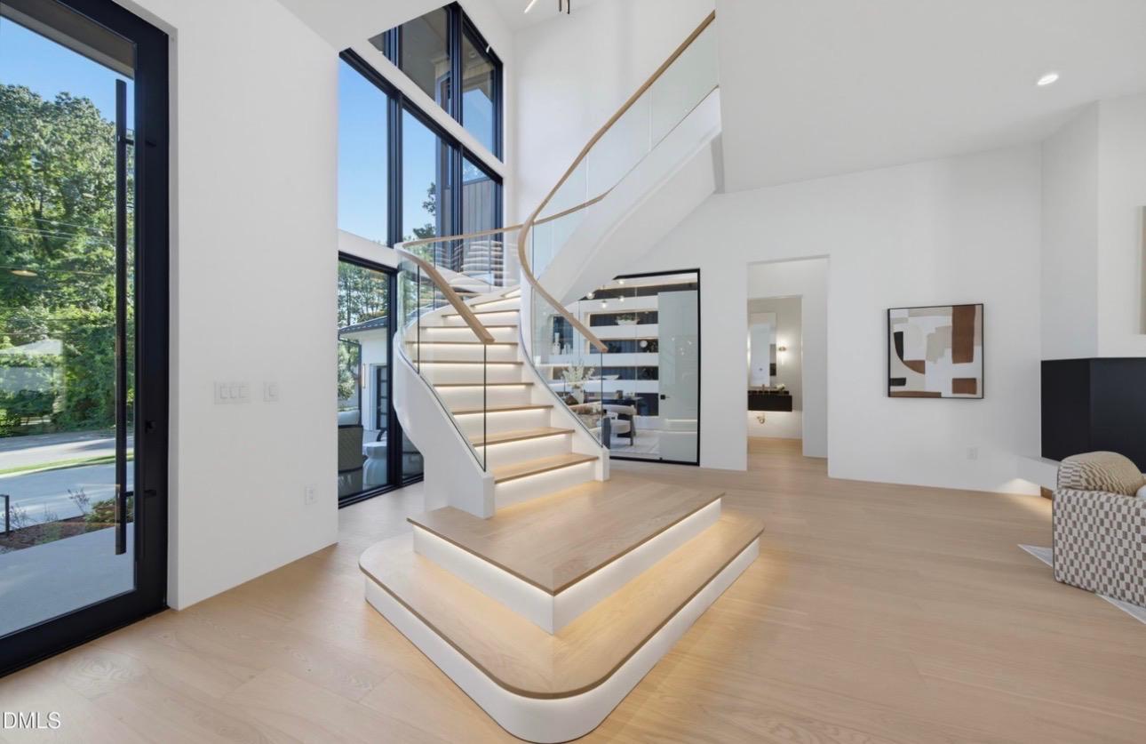 Bright modern luxury interior featuring an architectural curved floating staircase with integrated lighting, soaring double-height windows, and wide-plank white oak flooring