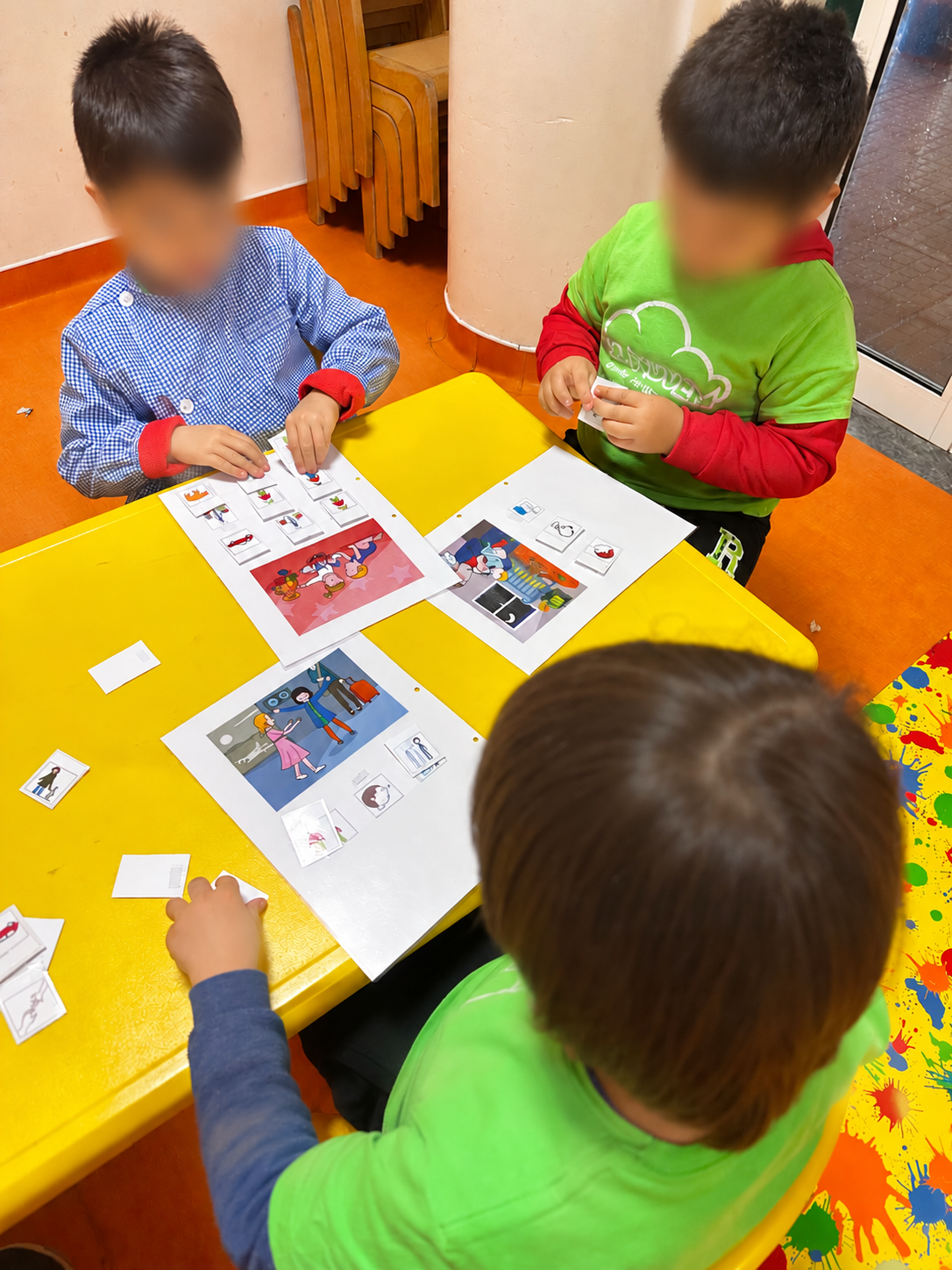 Crianças em mesa amarela com rostos protegidos a explorar materiais de construção frásica nas Oficinas de Estimulação da Linguagem