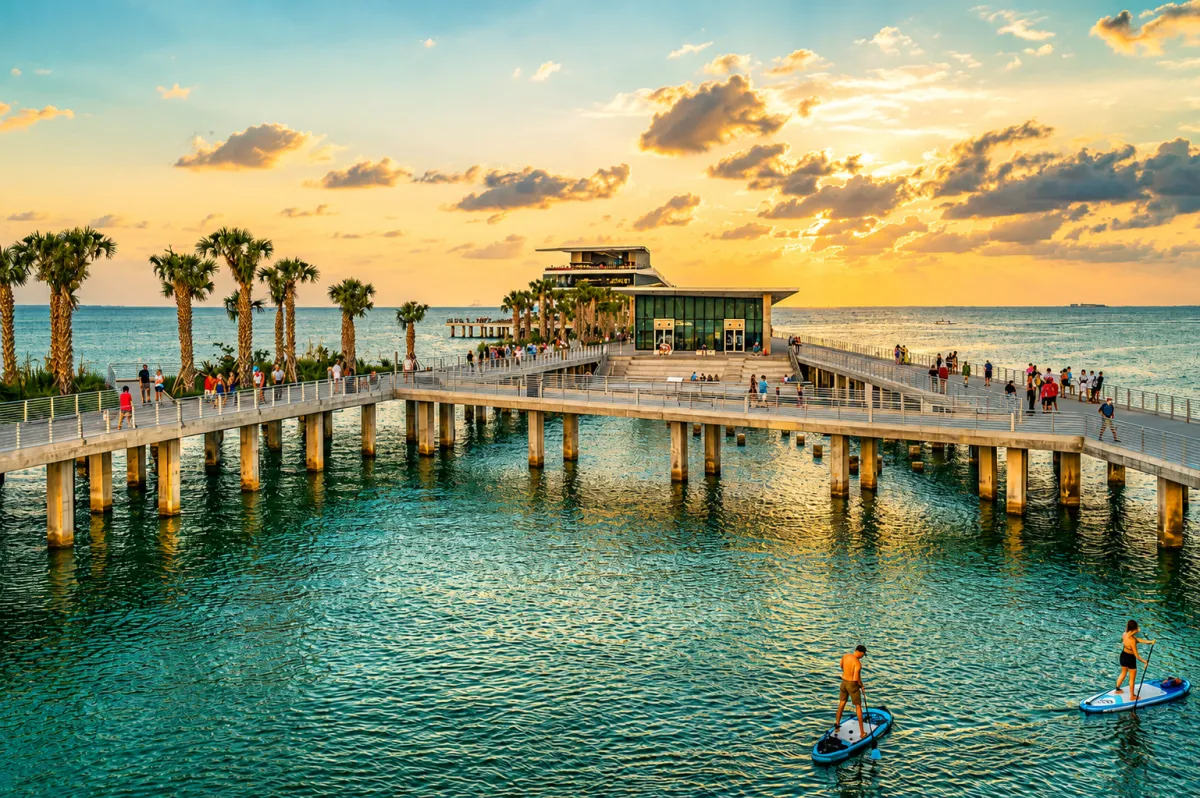 Paddleboarders in calm turquoise water by the St. Pete Pier promenade at sunset