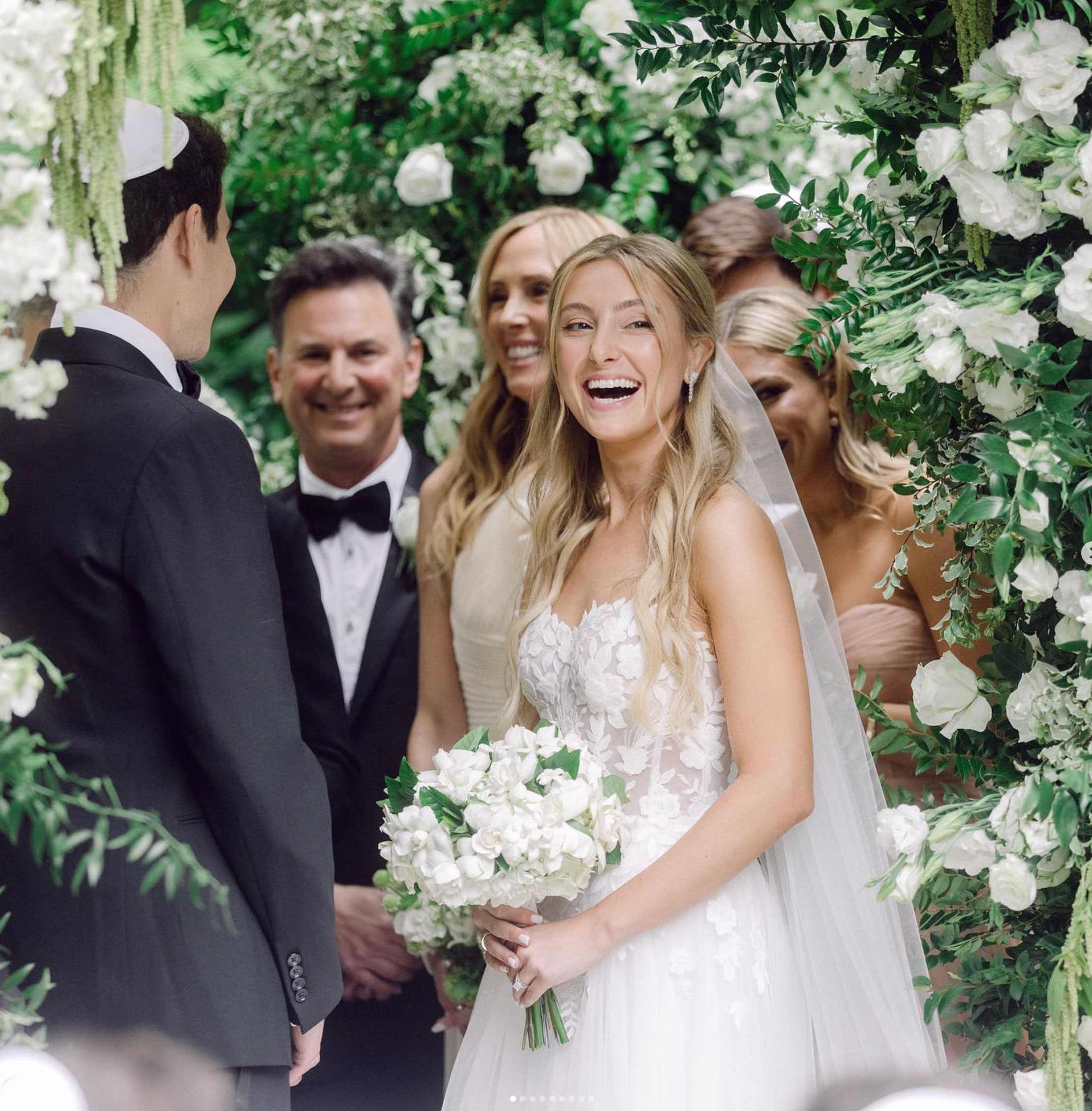 Joyful bride with white bouquet under a lush floral canopy designed by Creative Floral Designs