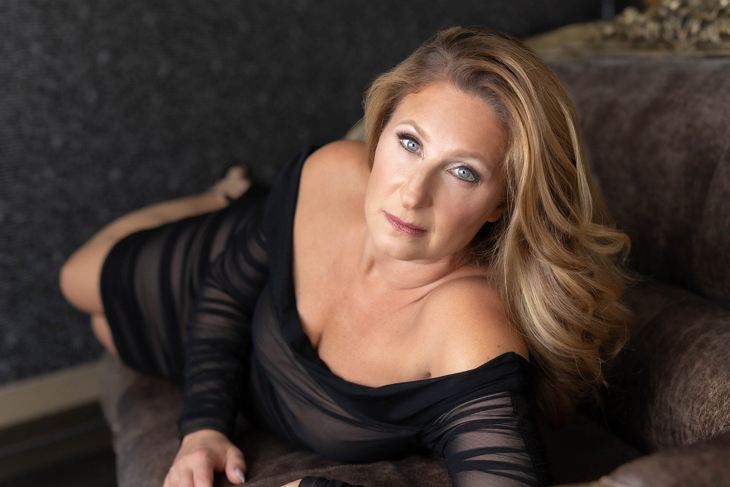 Boudoir portrait of a blonde woman in a sheer black dress reclining on a velvet chaise in the Jules Secret studio