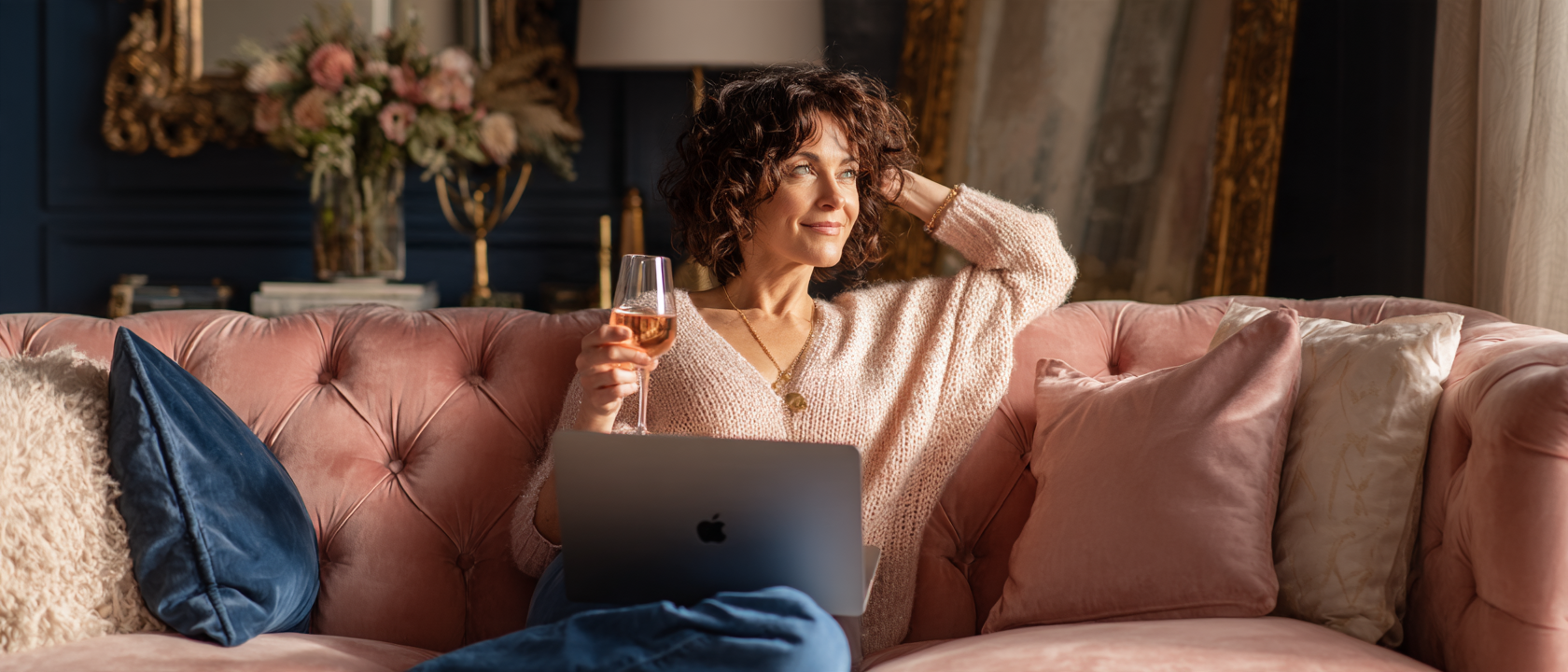 A woman relaxed on a dusty rose velvet couch holding a glass of wine with her laptop open — sovereign, easeful, and fully supported in her business