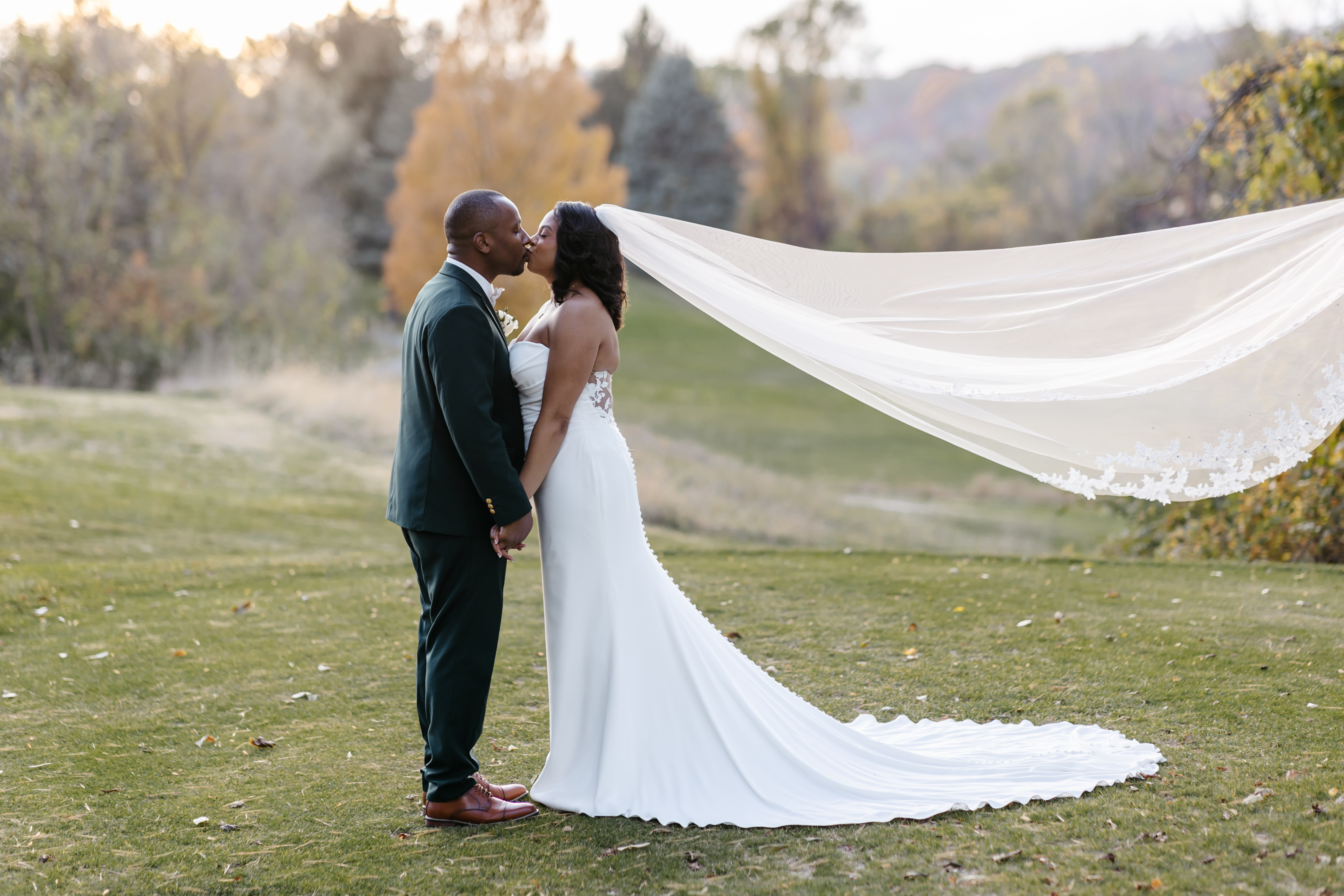 Bride and groom sharing a romantic kiss with dramatic veil caught in the breeze across scenic lawn