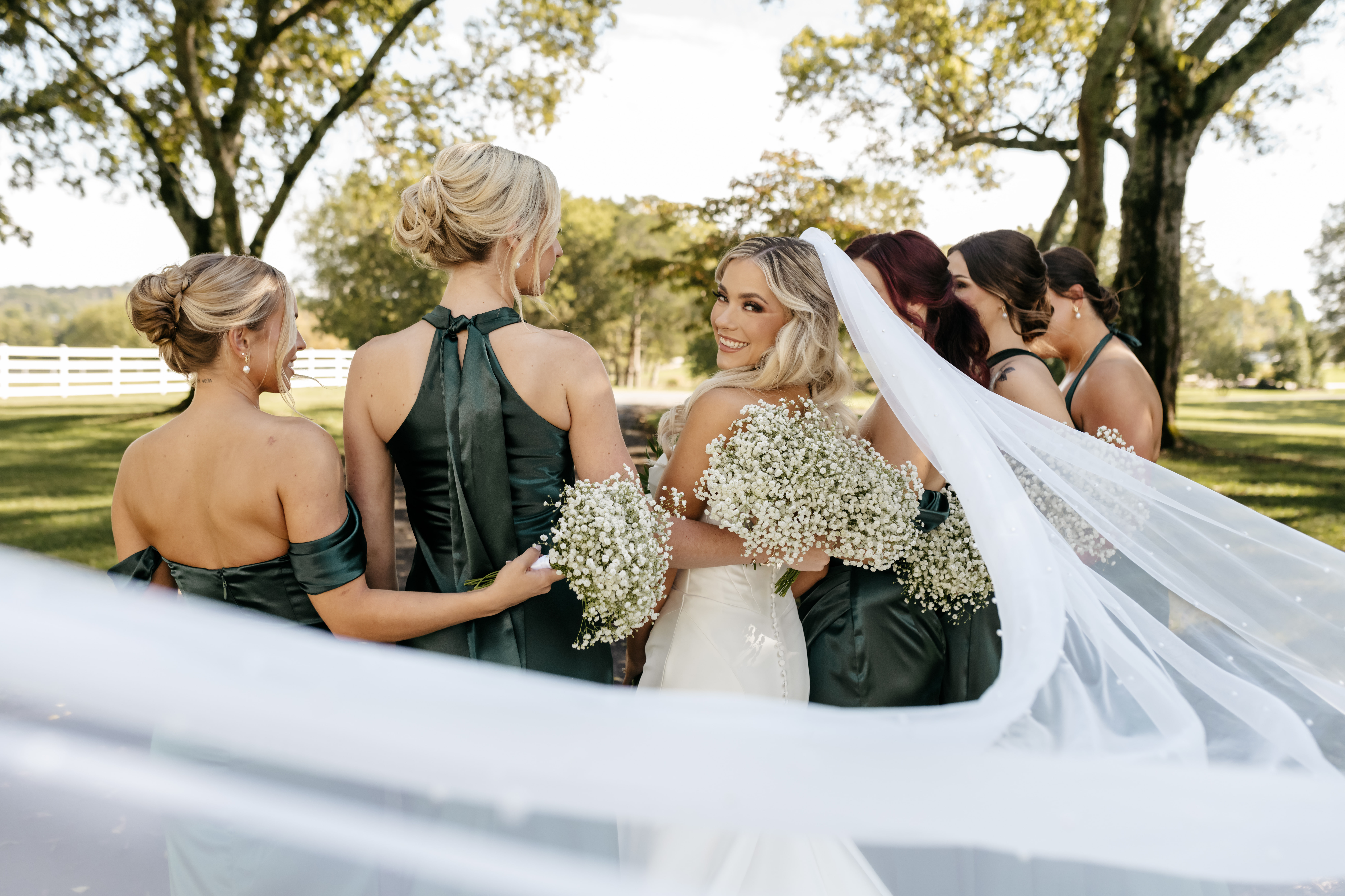 Bride smiling over shoulder surrounded by bridesmaids in emerald green gowns with baby's breath bouquets