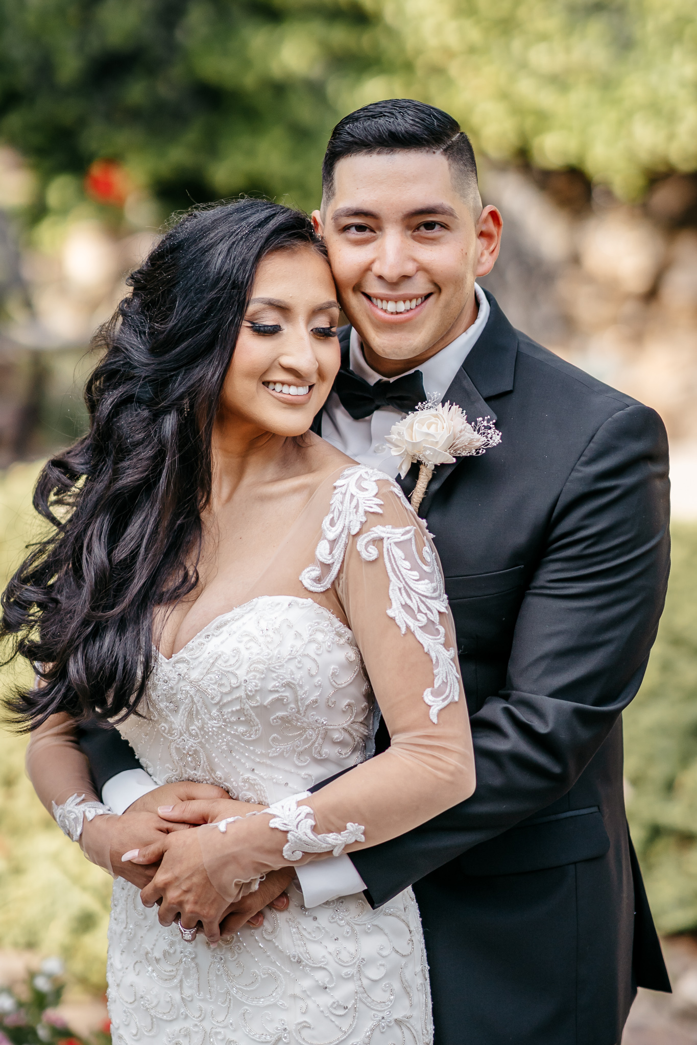Romantic close-up portrait of bride in embroidered sheer sleeve gown embraced by smiling groom in tuxedo