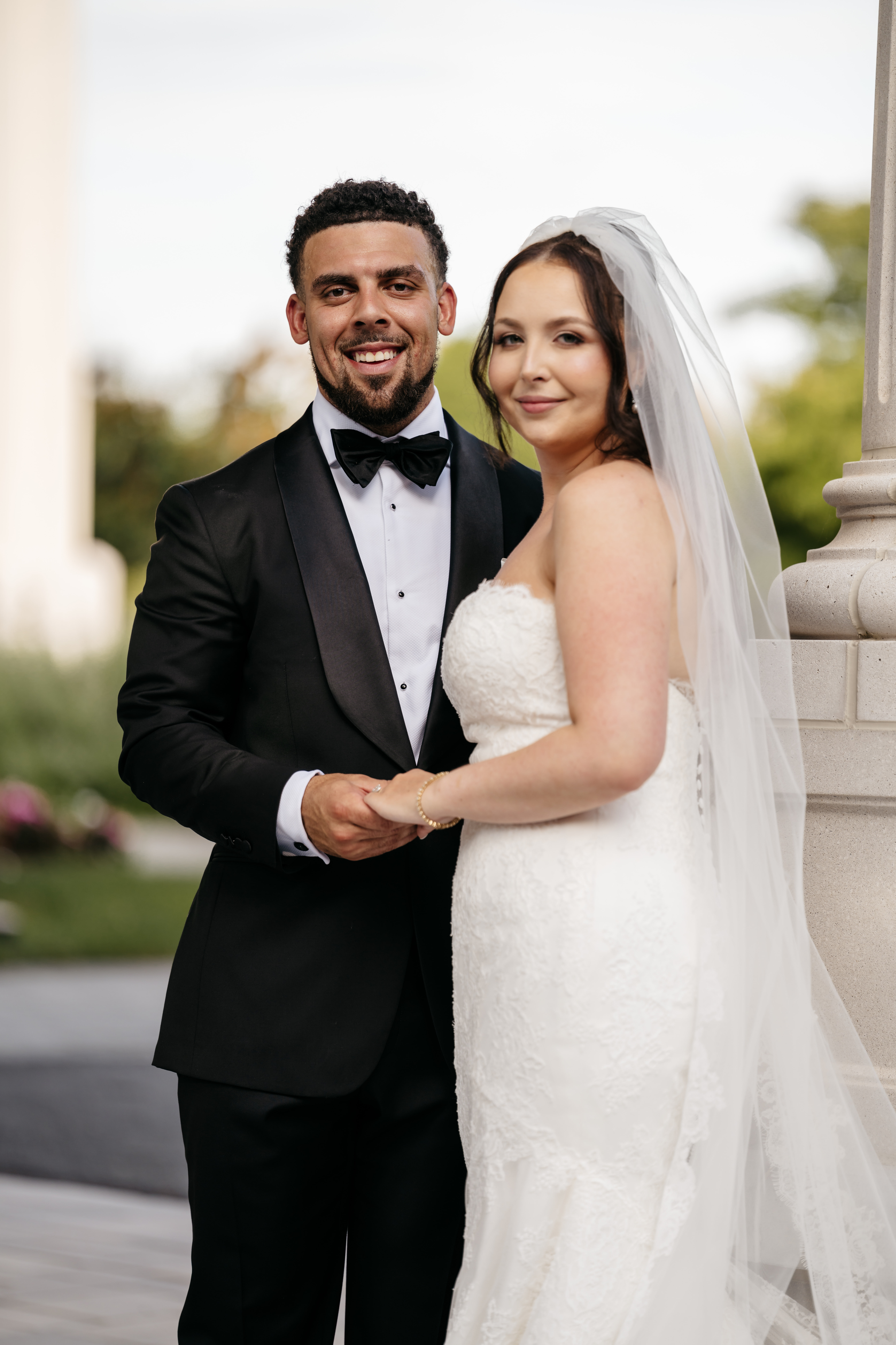 Bride looking over shoulder holding hands with groom along walkway lined with blooming red roses