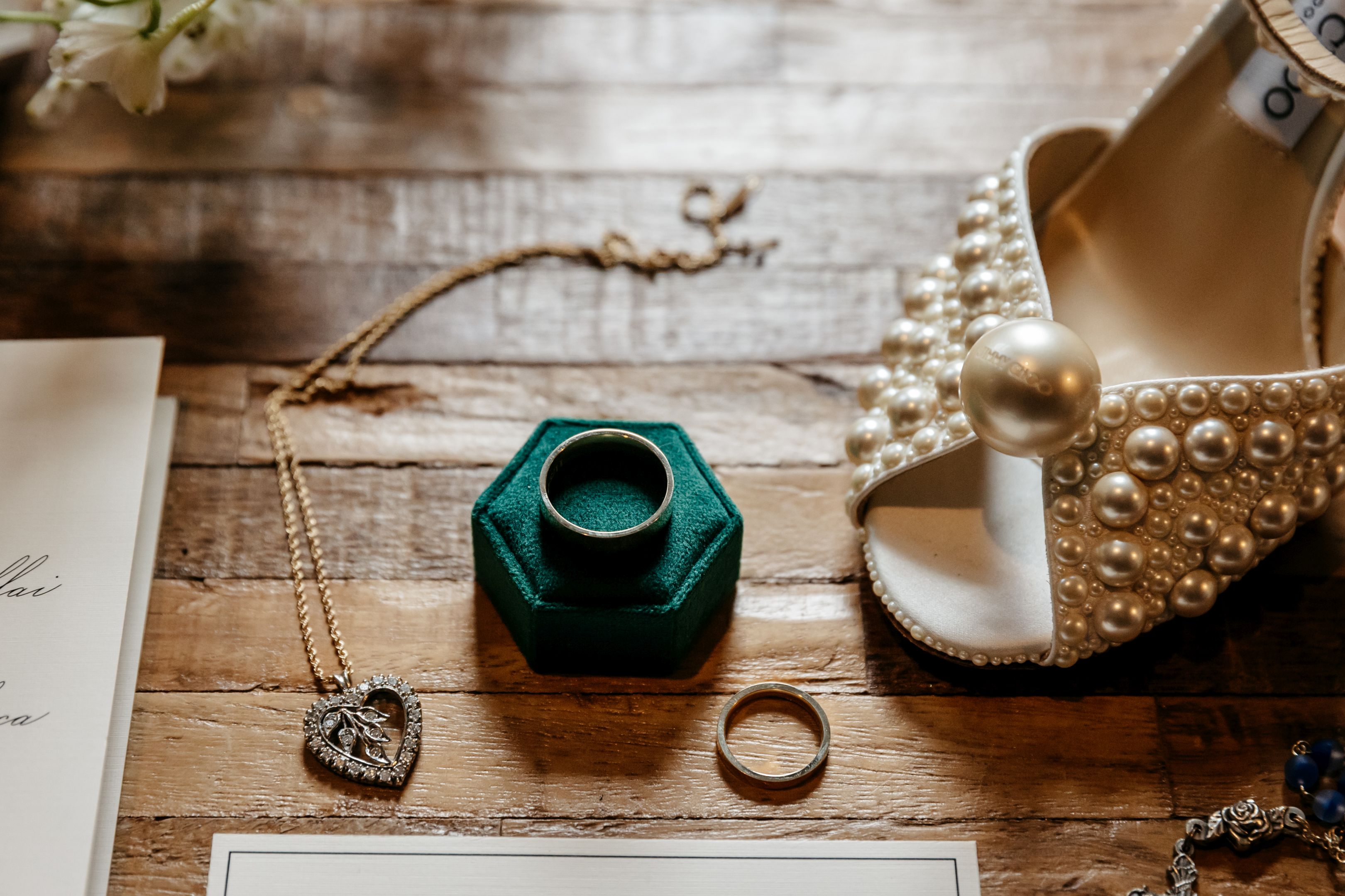 Bridal details flat lay with pearl heel shoe, emerald green velvet ring box, wedding rings, and heart pendant necklace