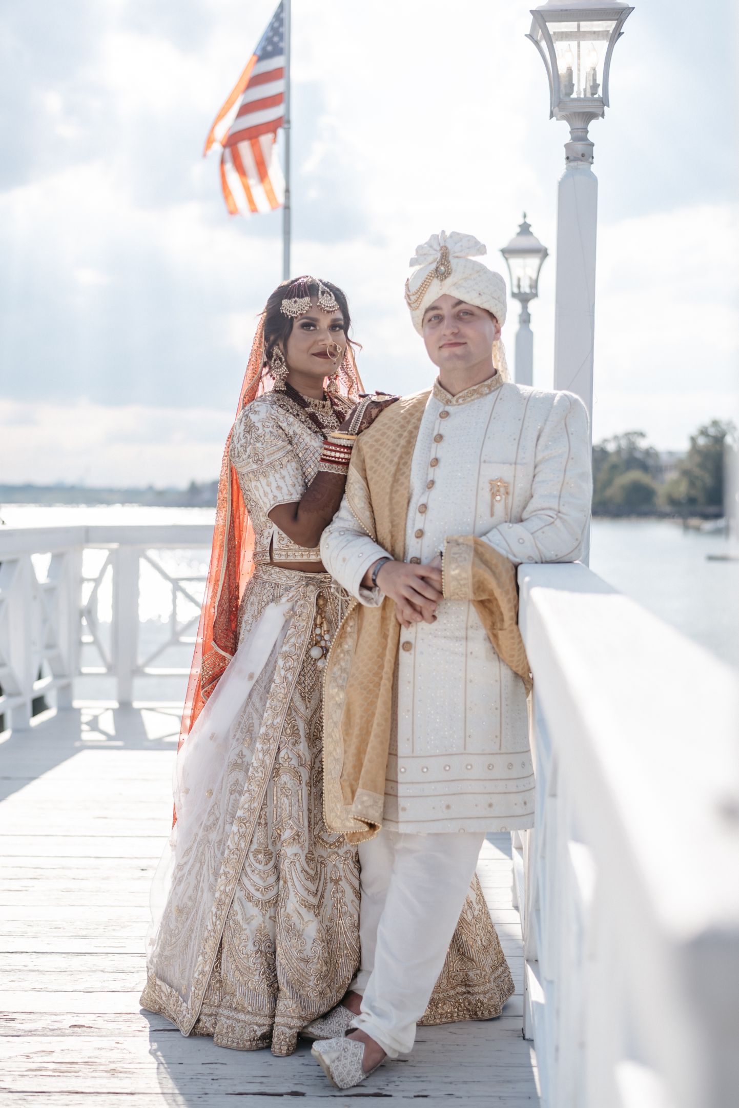 Bride and groom in ornate ivory and gold sherwani and lehenga on sunlit wooden pier