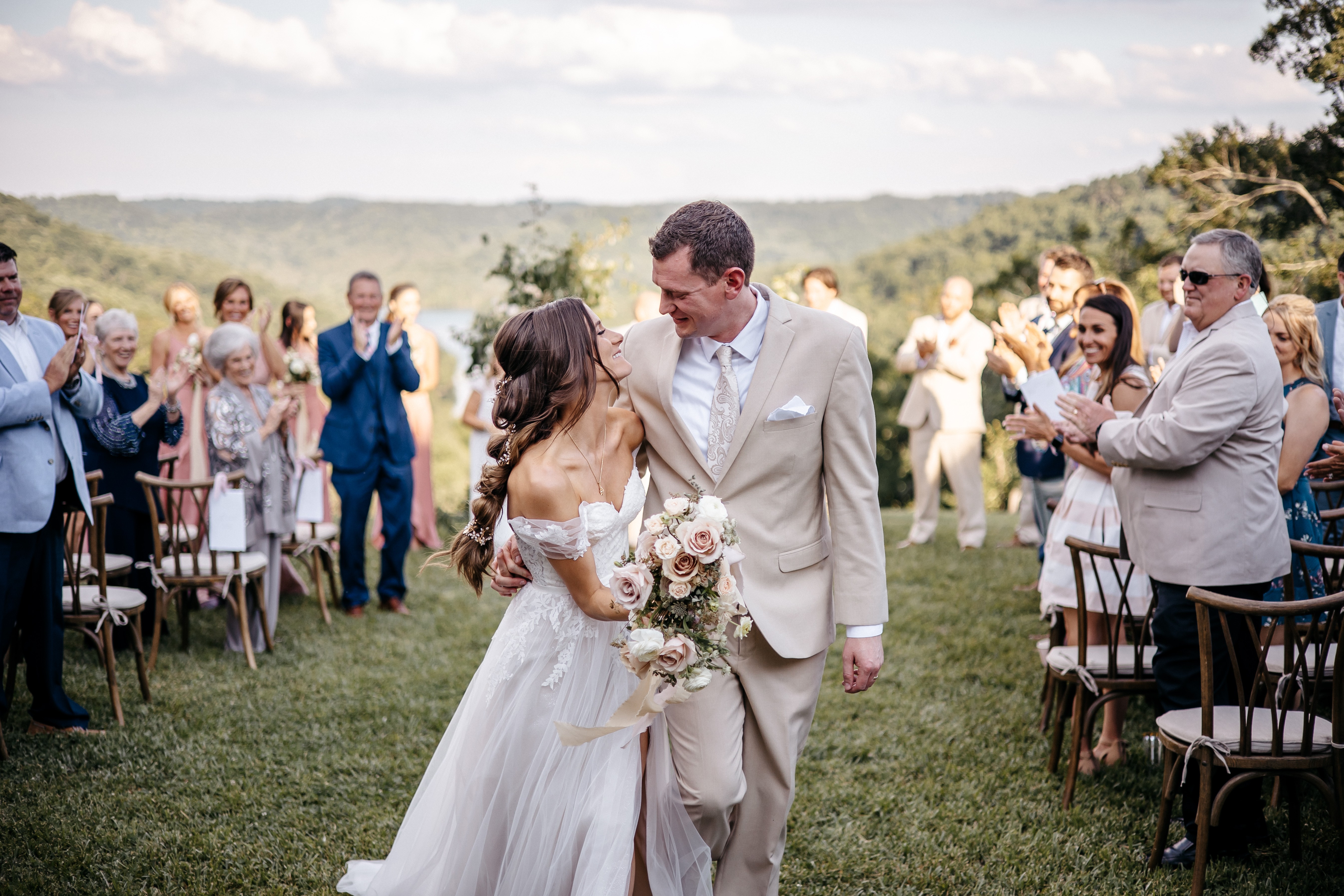 Joyful bride and groom smiling and walking down outdoor ceremony aisle with guests applauding