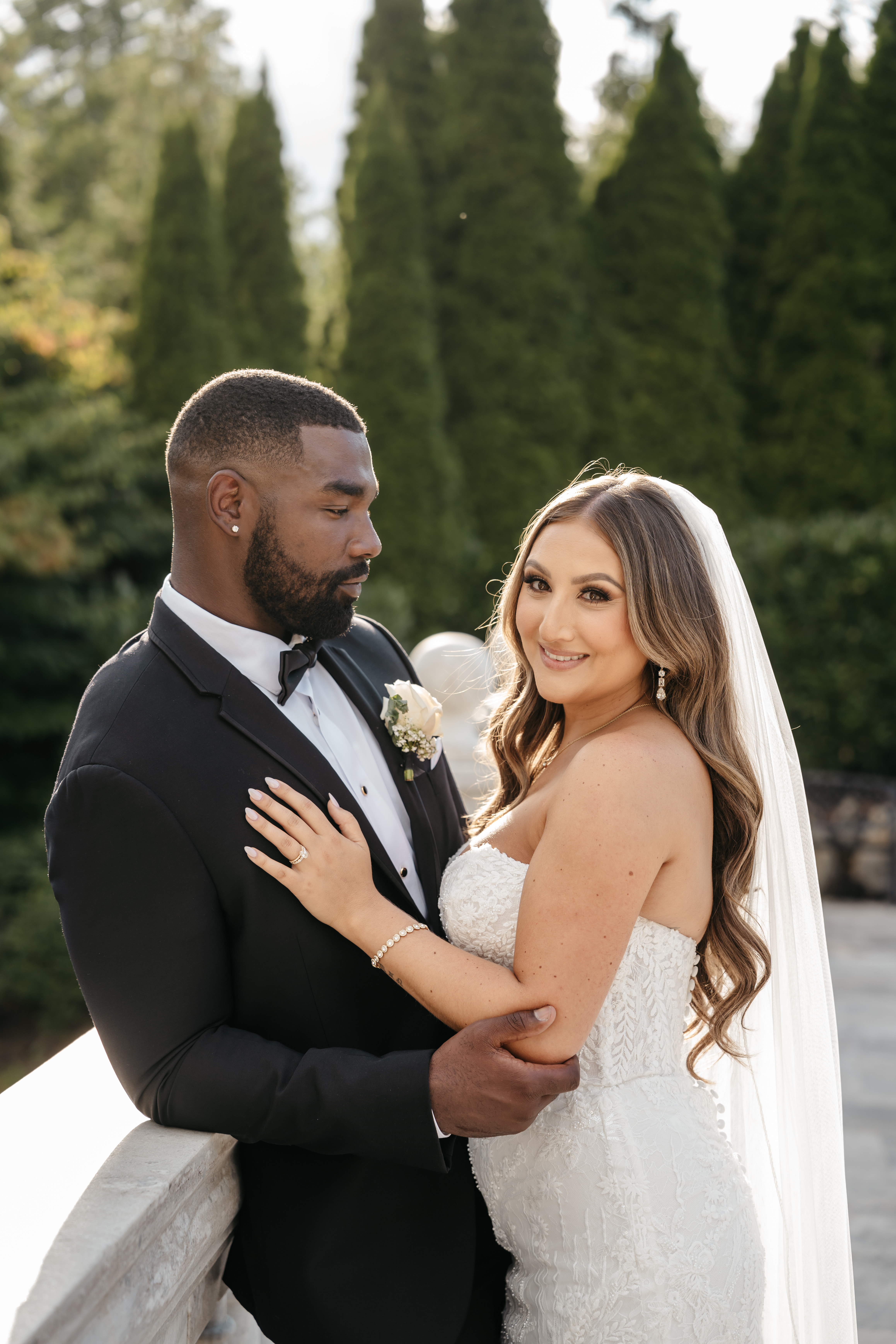 Bride with long flowing hair and groom leaning on stone balustrade in front of lush evergreen pines