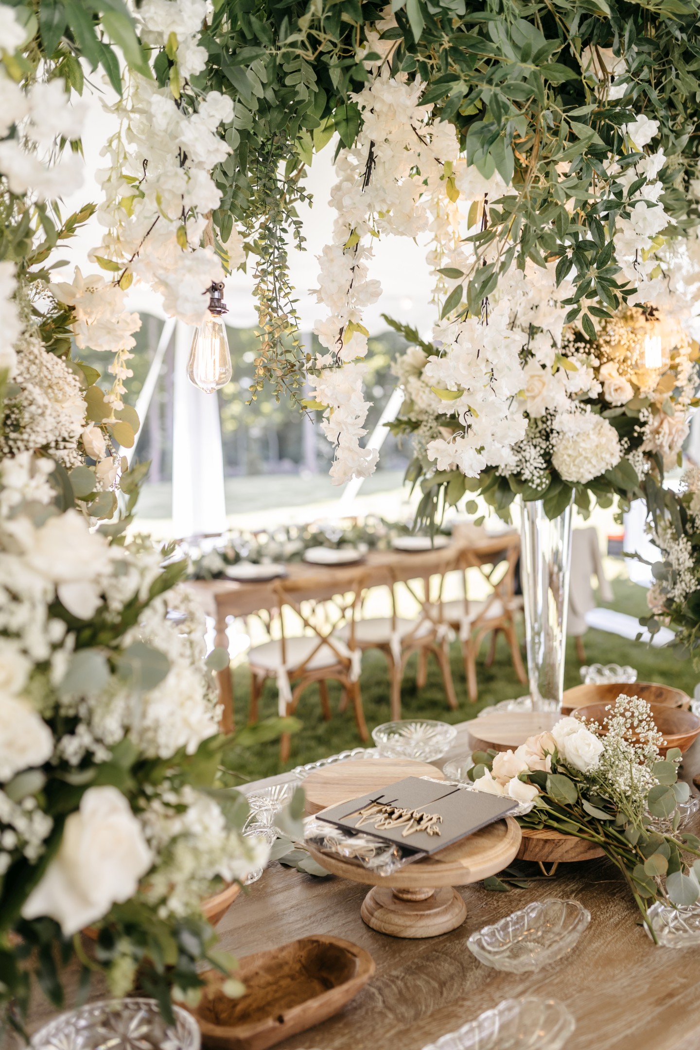 Lush wedding reception tent with hanging white florals, Edison bulb, and elegant wooden tablescape