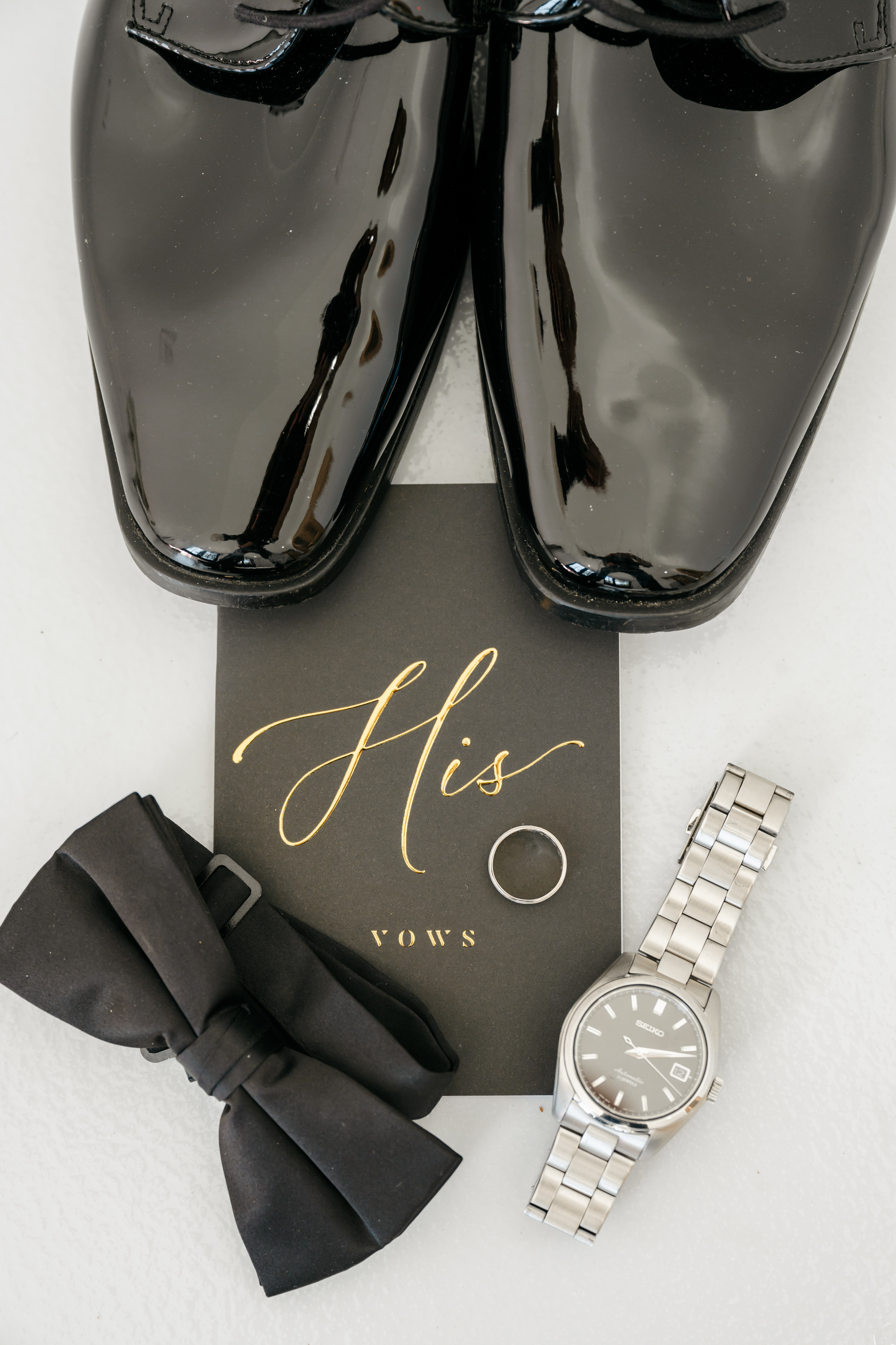 Flat lay of groom accessories with black dress shoes, His Vows booklet, wedding ring, and watch