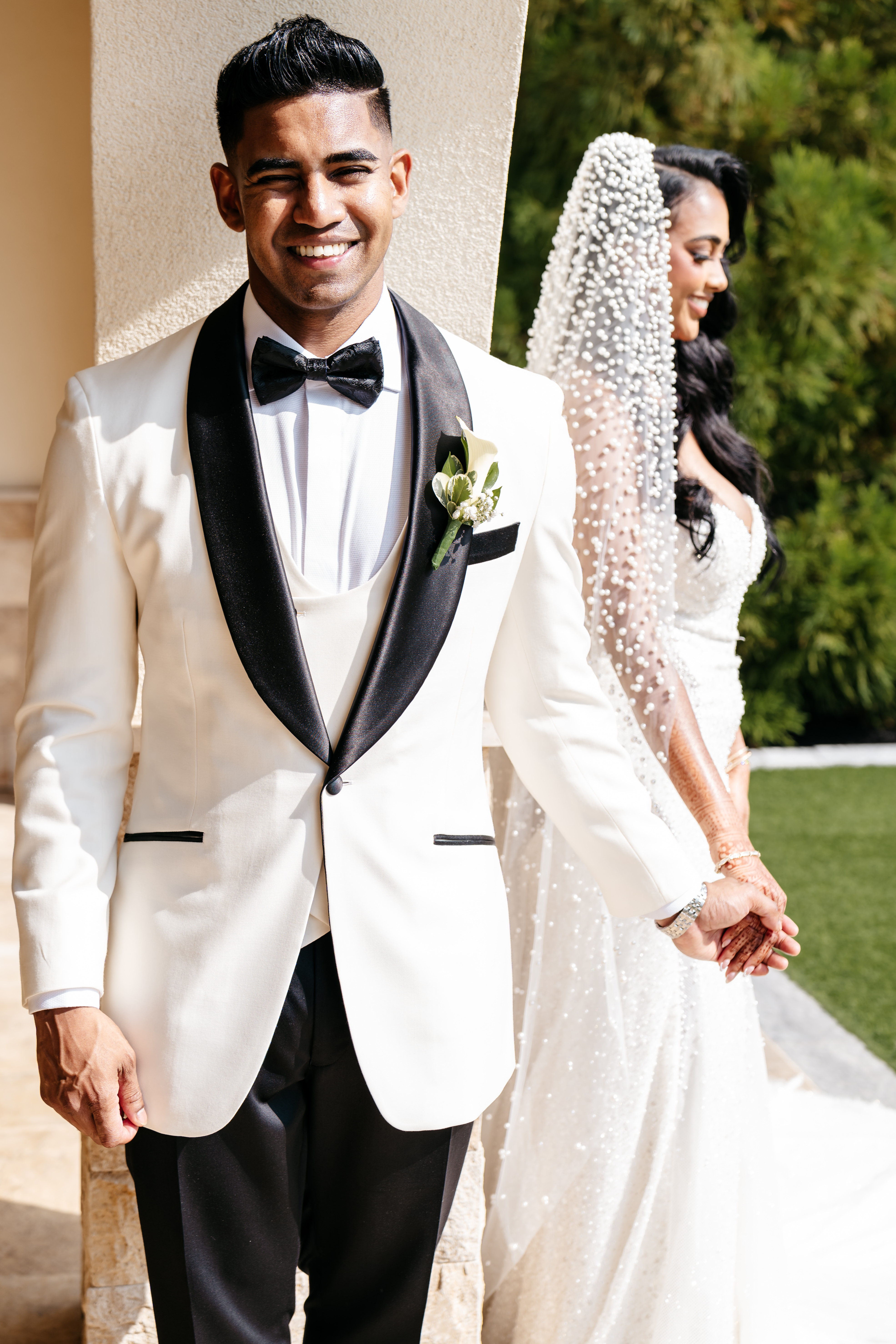 Smiling groom in ivory tuxedo holding hands with bride in pearl-studded veil