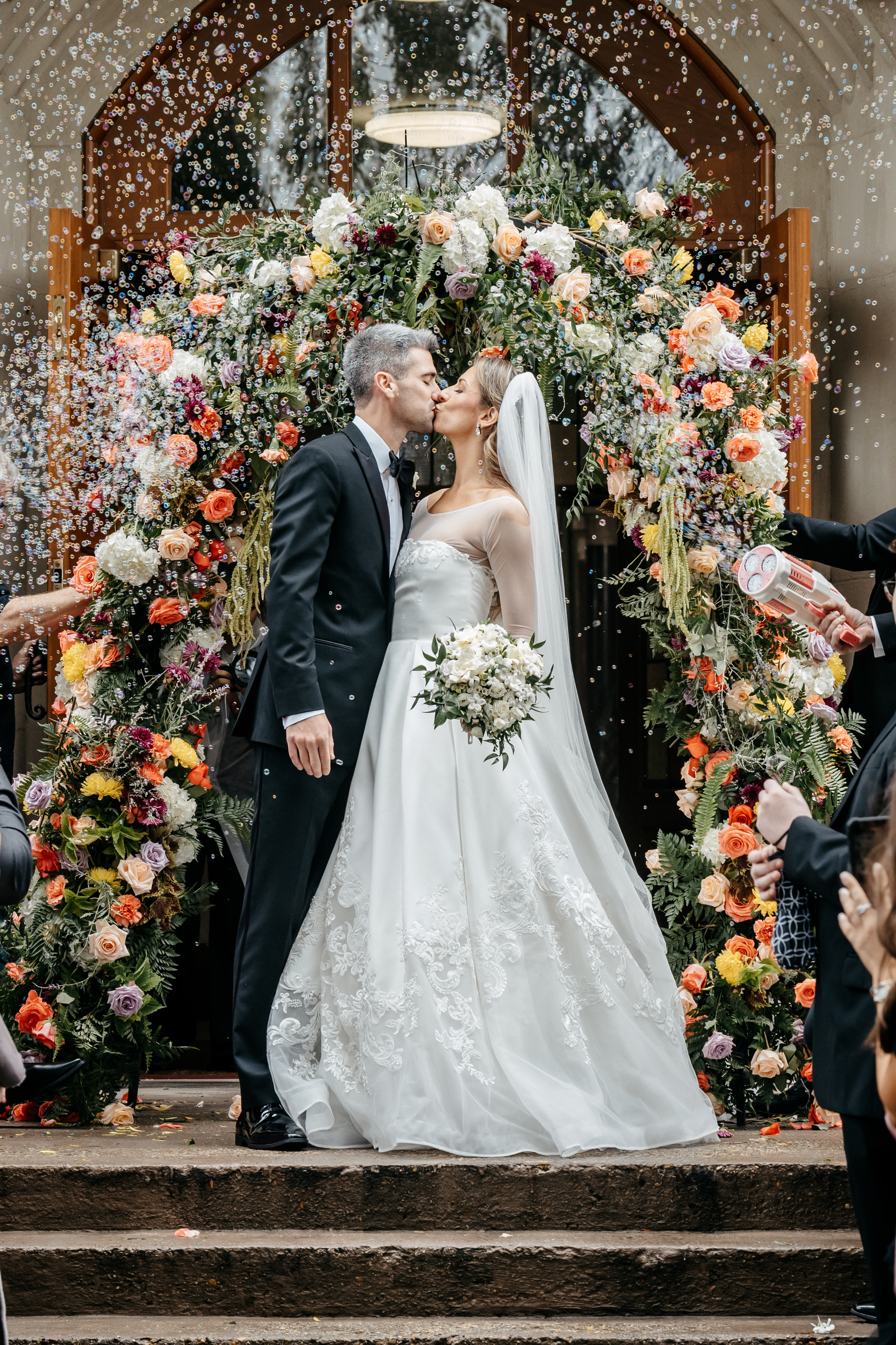 Bride and groom sharing a tender kiss under an elaborate floral arch surrounded by floating bubbles