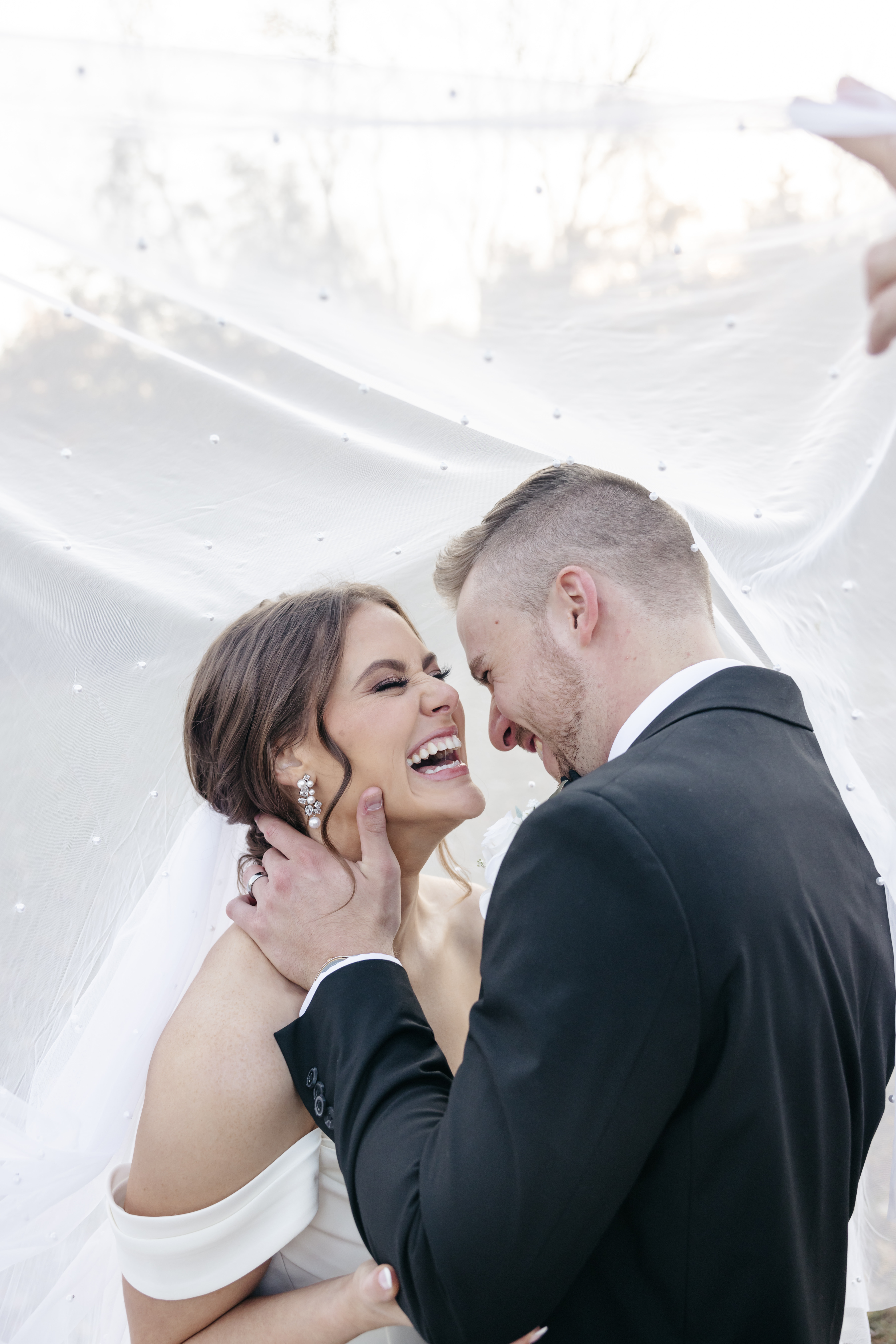 Bride and groom sharing an ecstatic laugh embracing under a pearl-dotted bridal veil