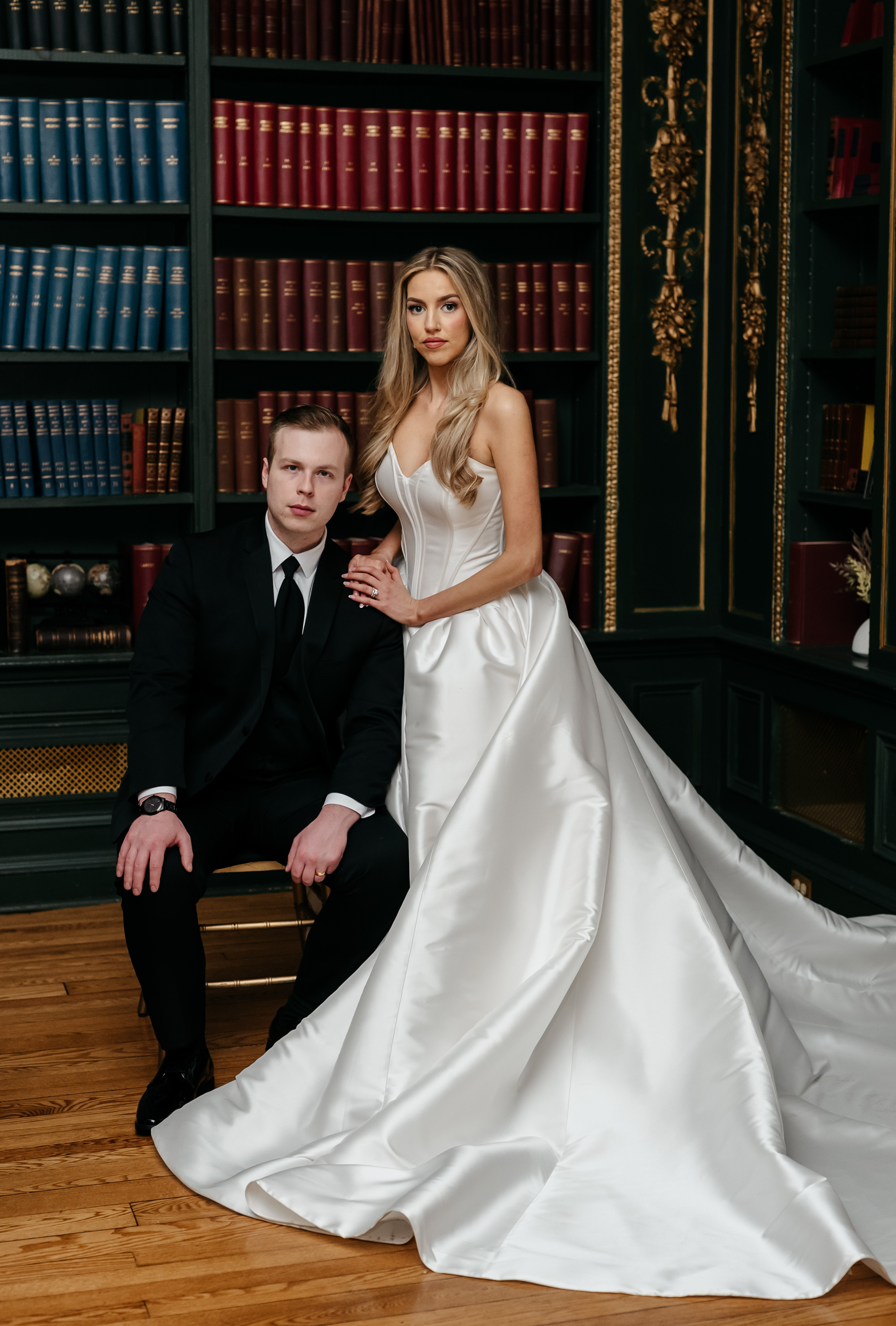 Bride in strapless ballgown with dramatic train beside seated groom in classic dark wood library