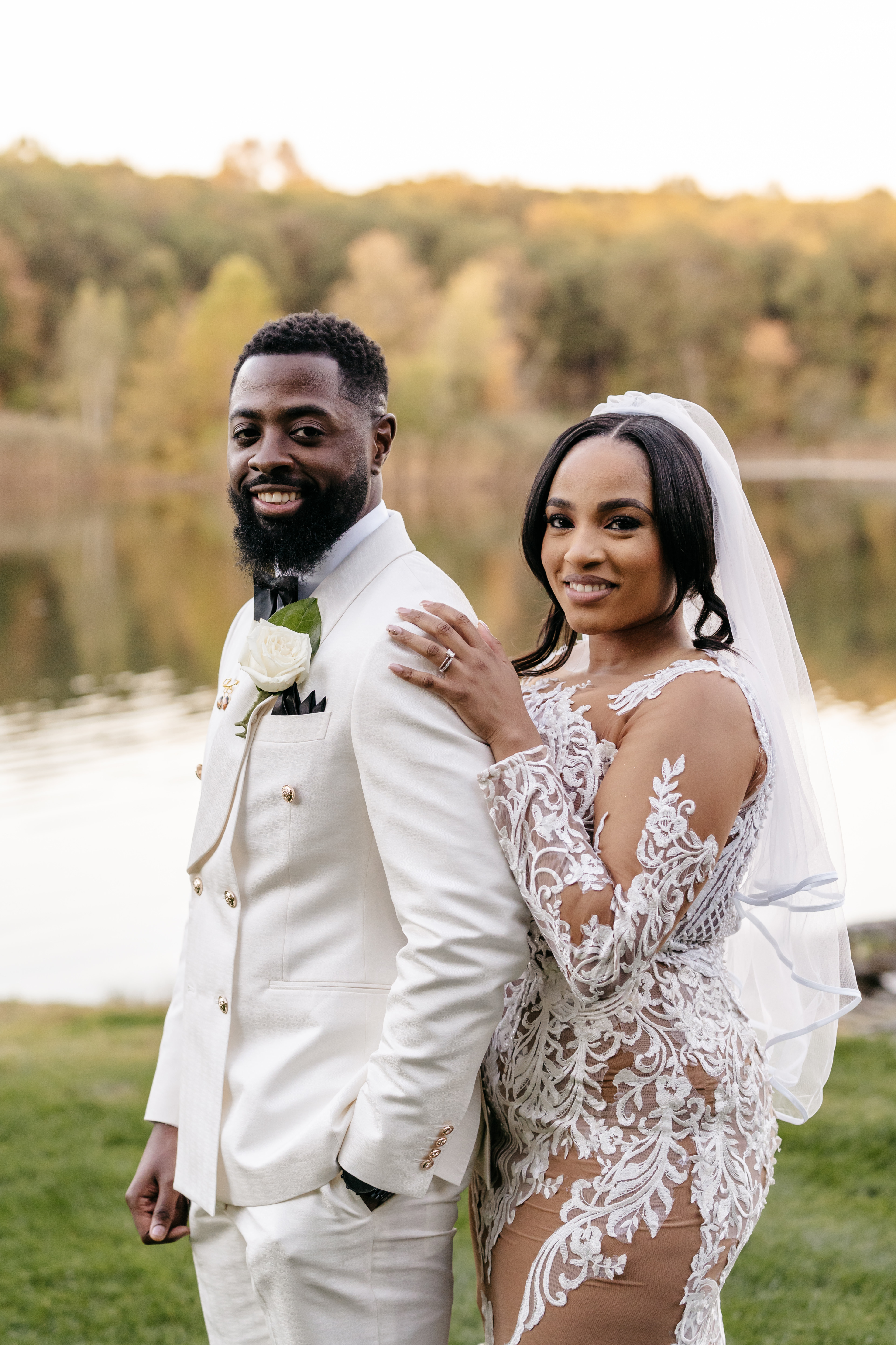 Smiling bride in lace gown and groom in cream double-breasted suit beside a tranquil lake
