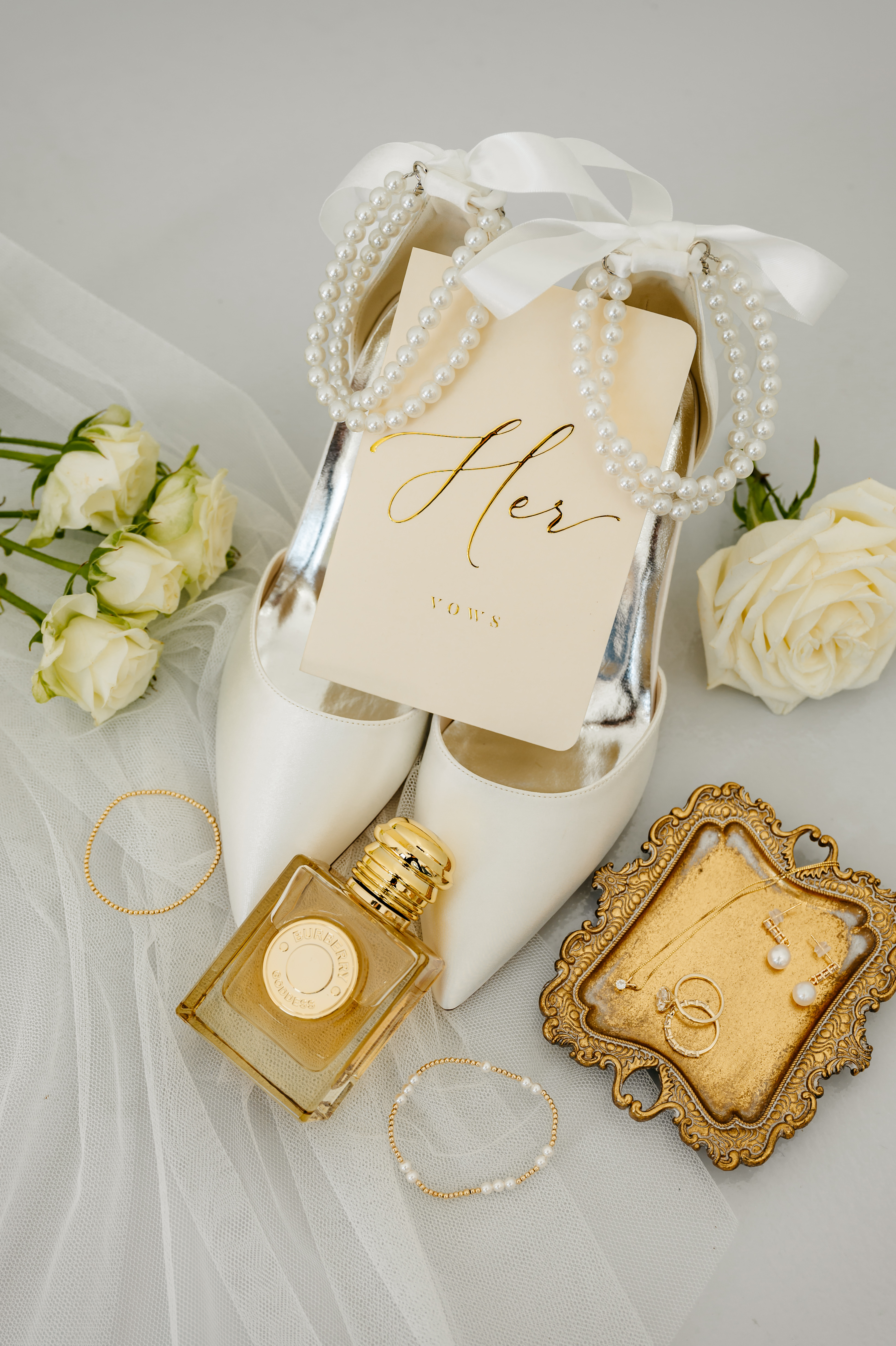 Flat lay of bridal details with pearl heels, Her Vows booklet, perfume, jewelry, and white roses
