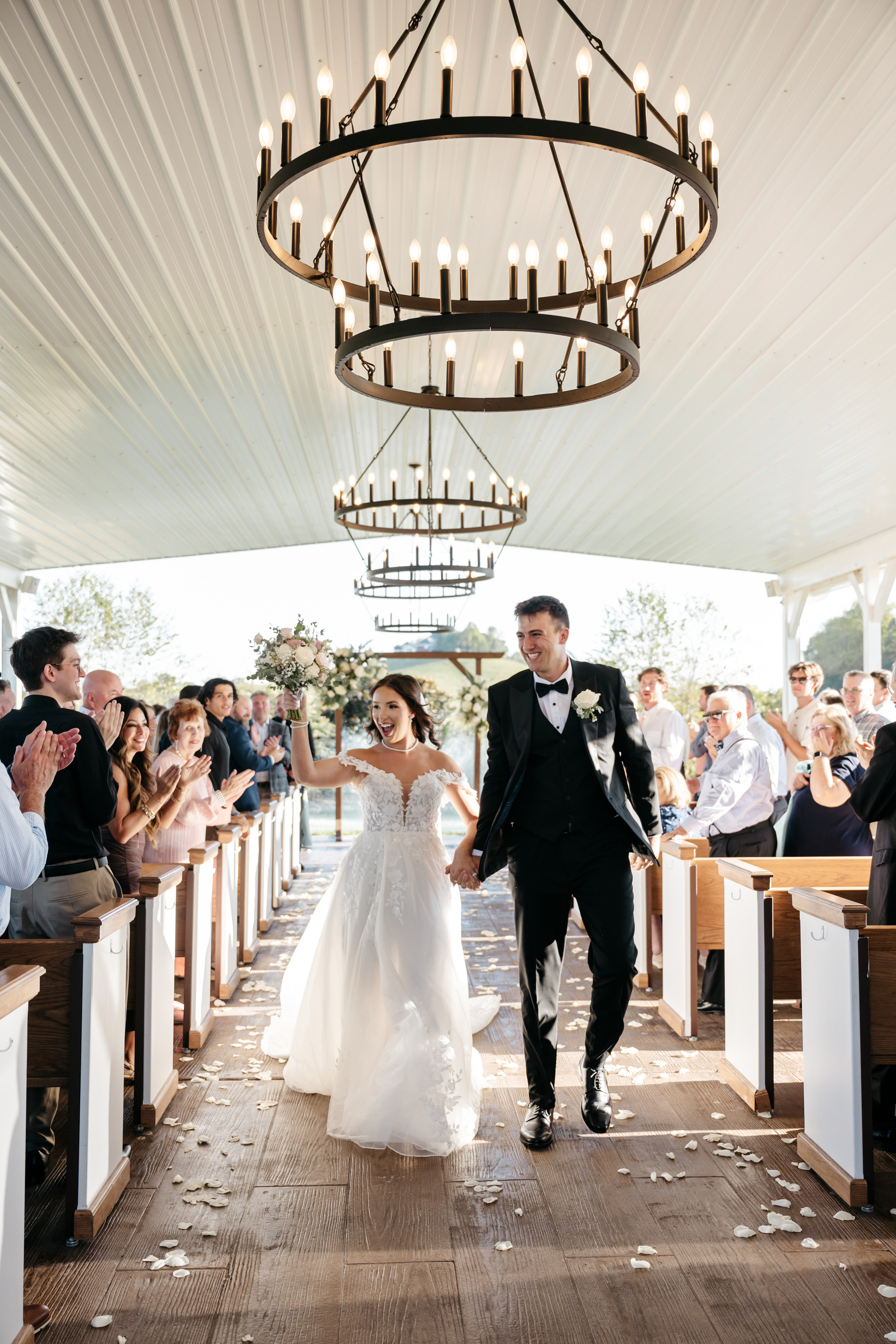 Joyful bride and groom celebrating down the covered outdoor chapel aisle under iron chandeliers
