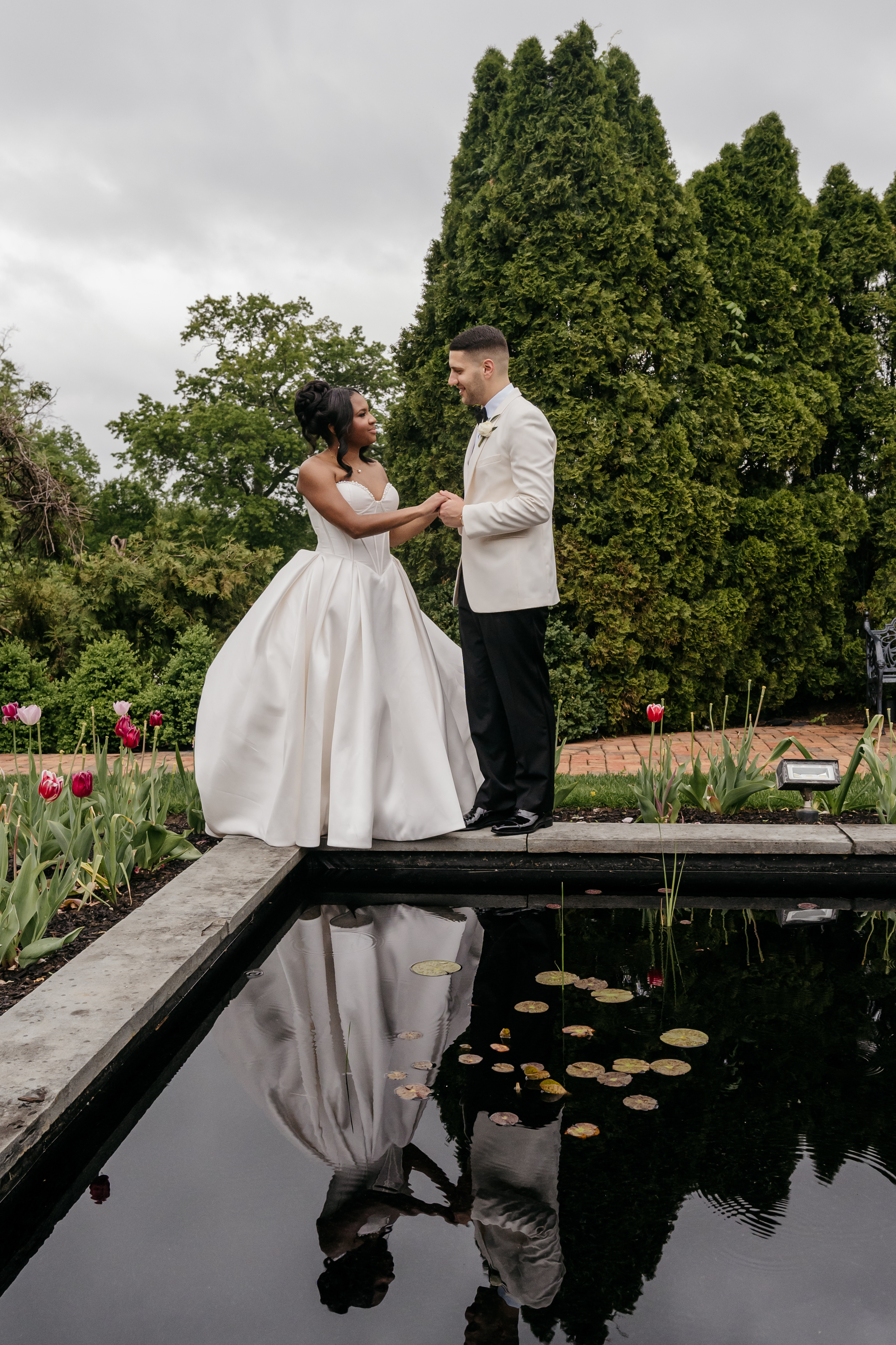 Bride in full satin ballgown and groom holding hands reflected in garden lily pond