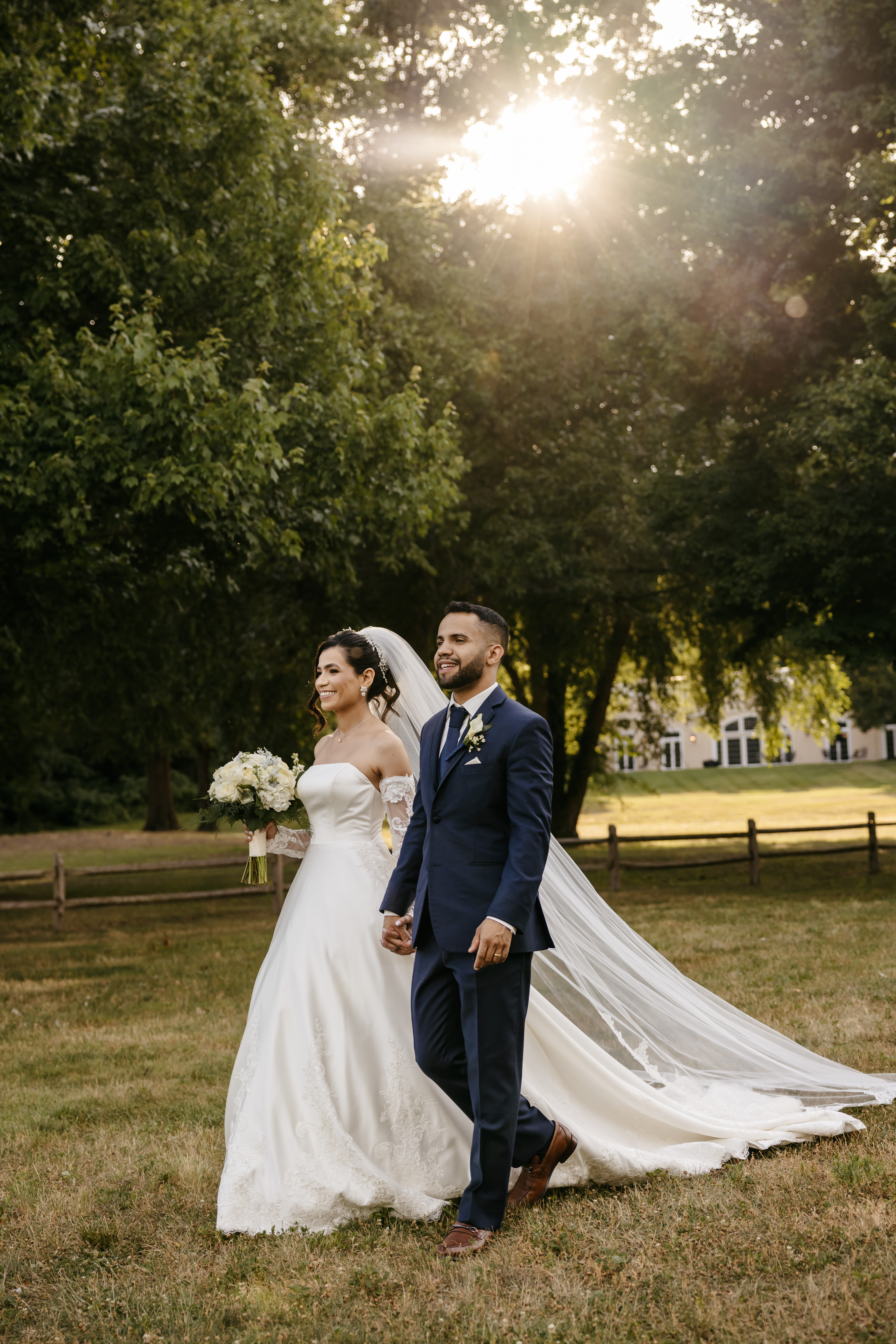Bride and groom walking in sunlit park with cathedral veil