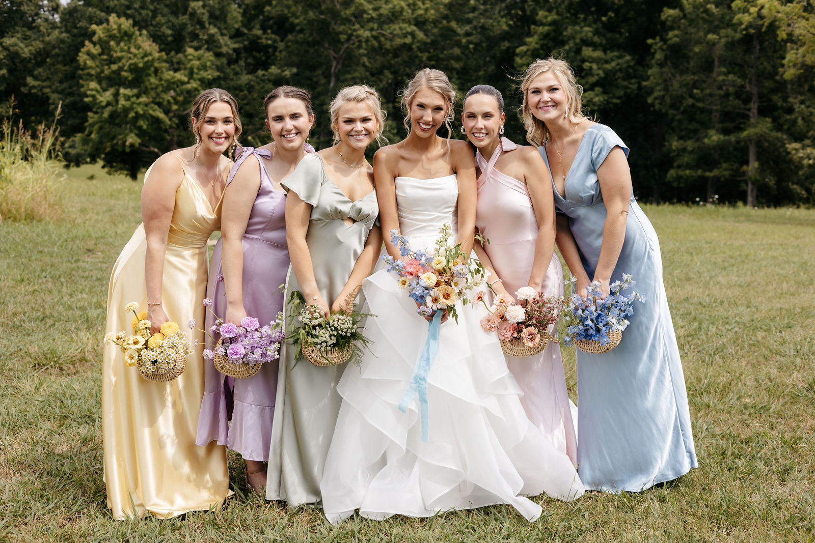Smiling bride in tiered gown surrounded by bridesmaids in pastel satin dresses holding floral baskets outdoors