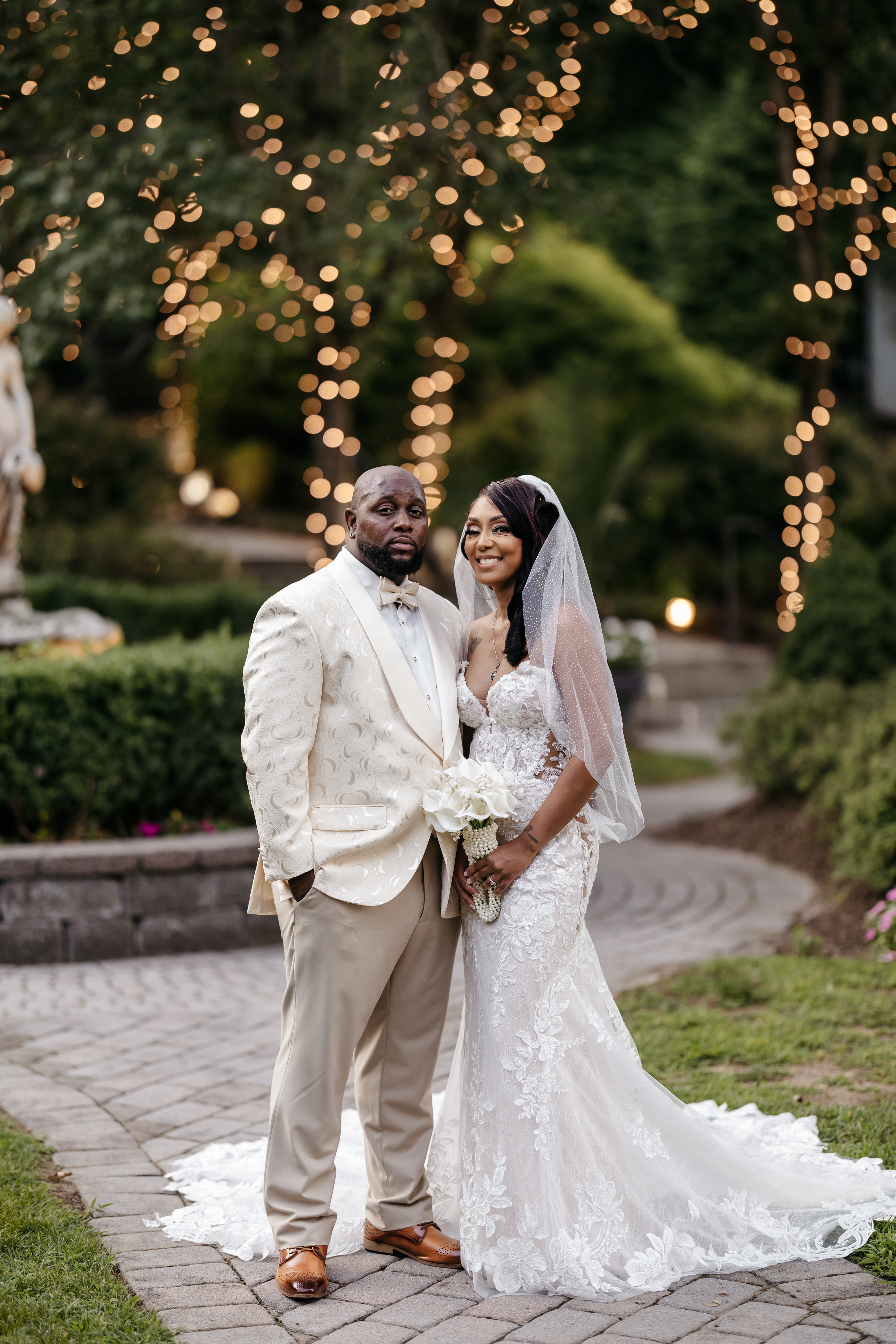 Bride in lace fitted gown and groom in champagne patterned tuxedo standing on stone path illuminated by string lights