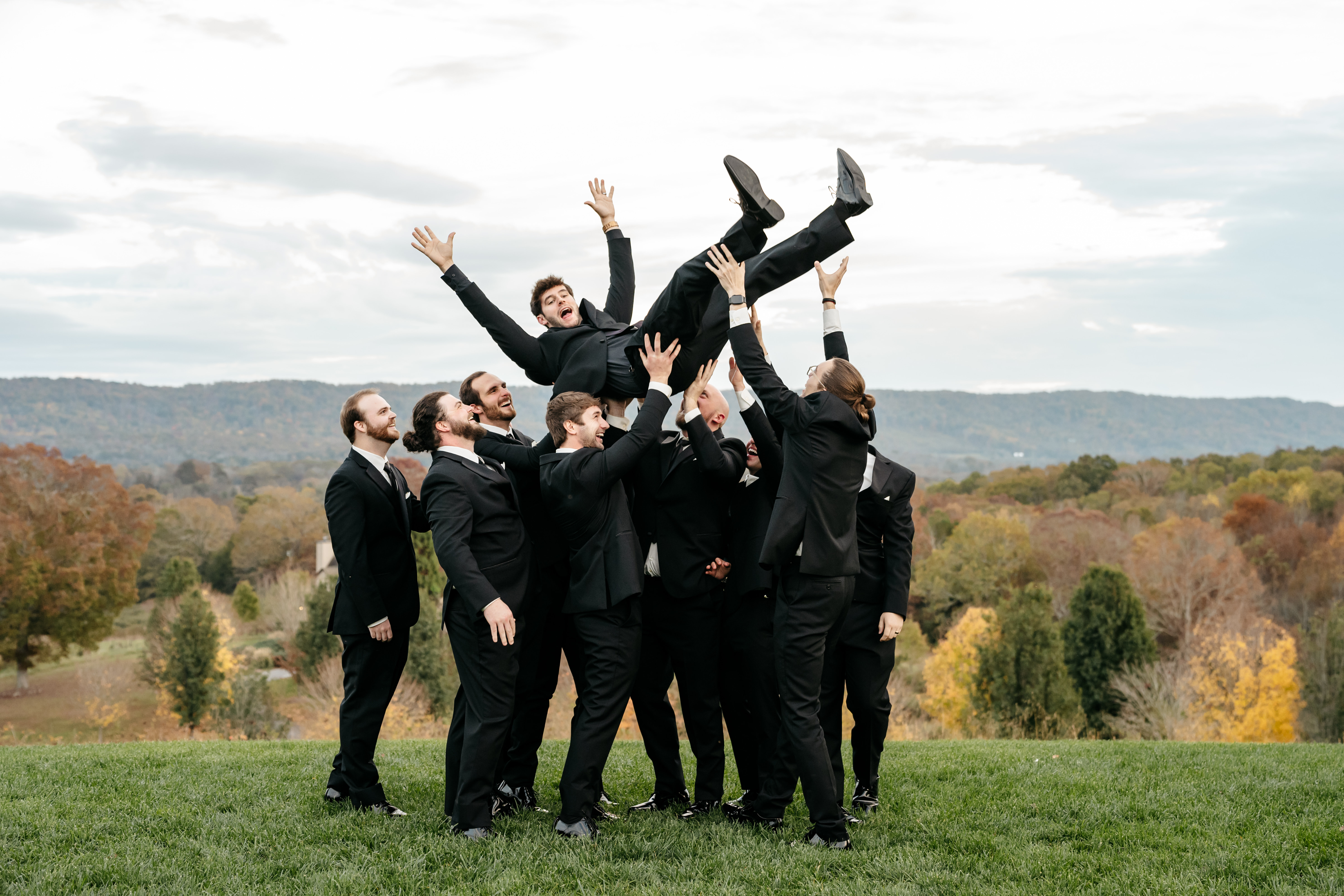 Groomsmen joyfully tossing the groom into the air on scenic hilltop lawn