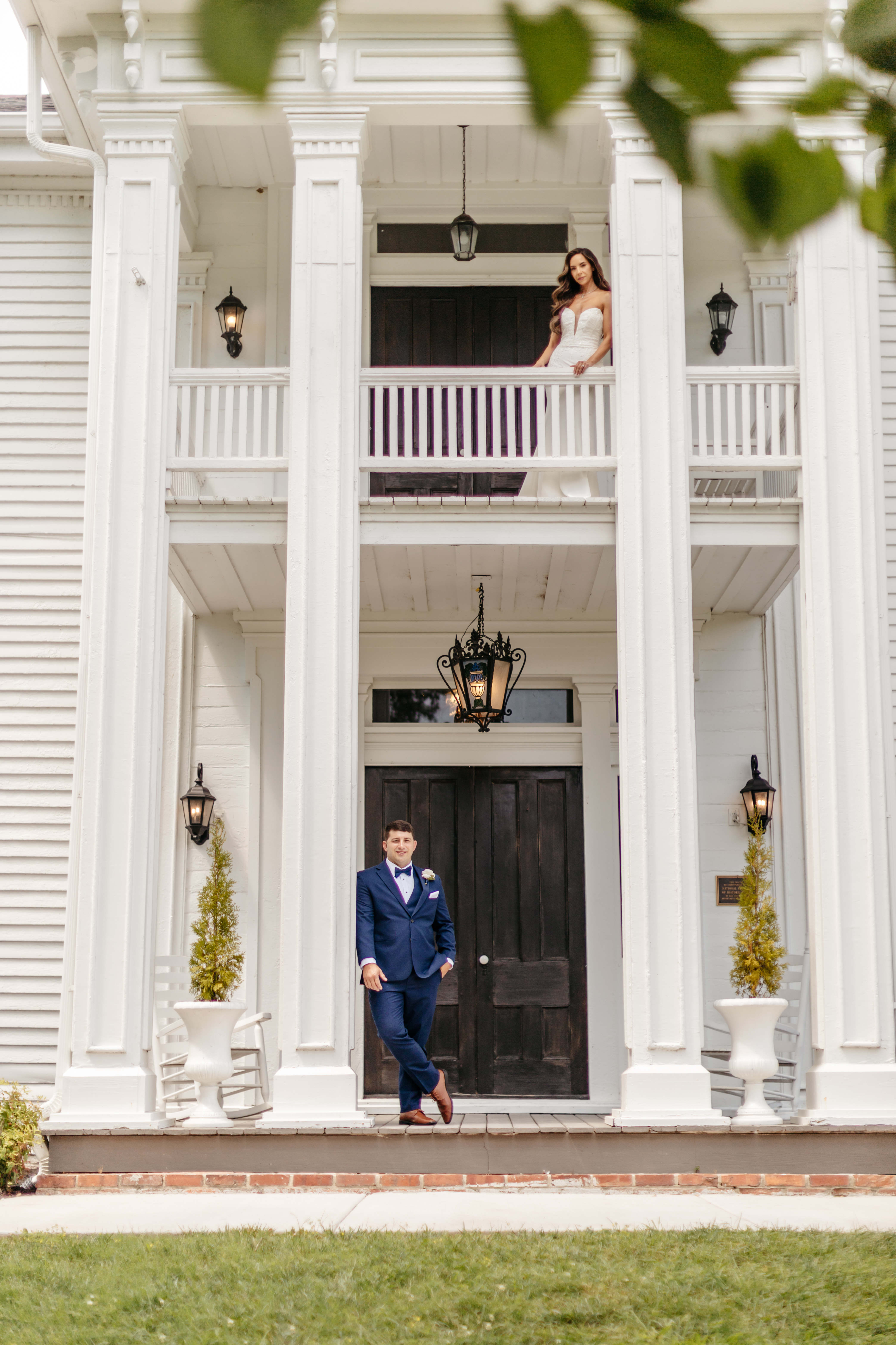 Groom in navy suit on grand white porch with bride on second-story balcony