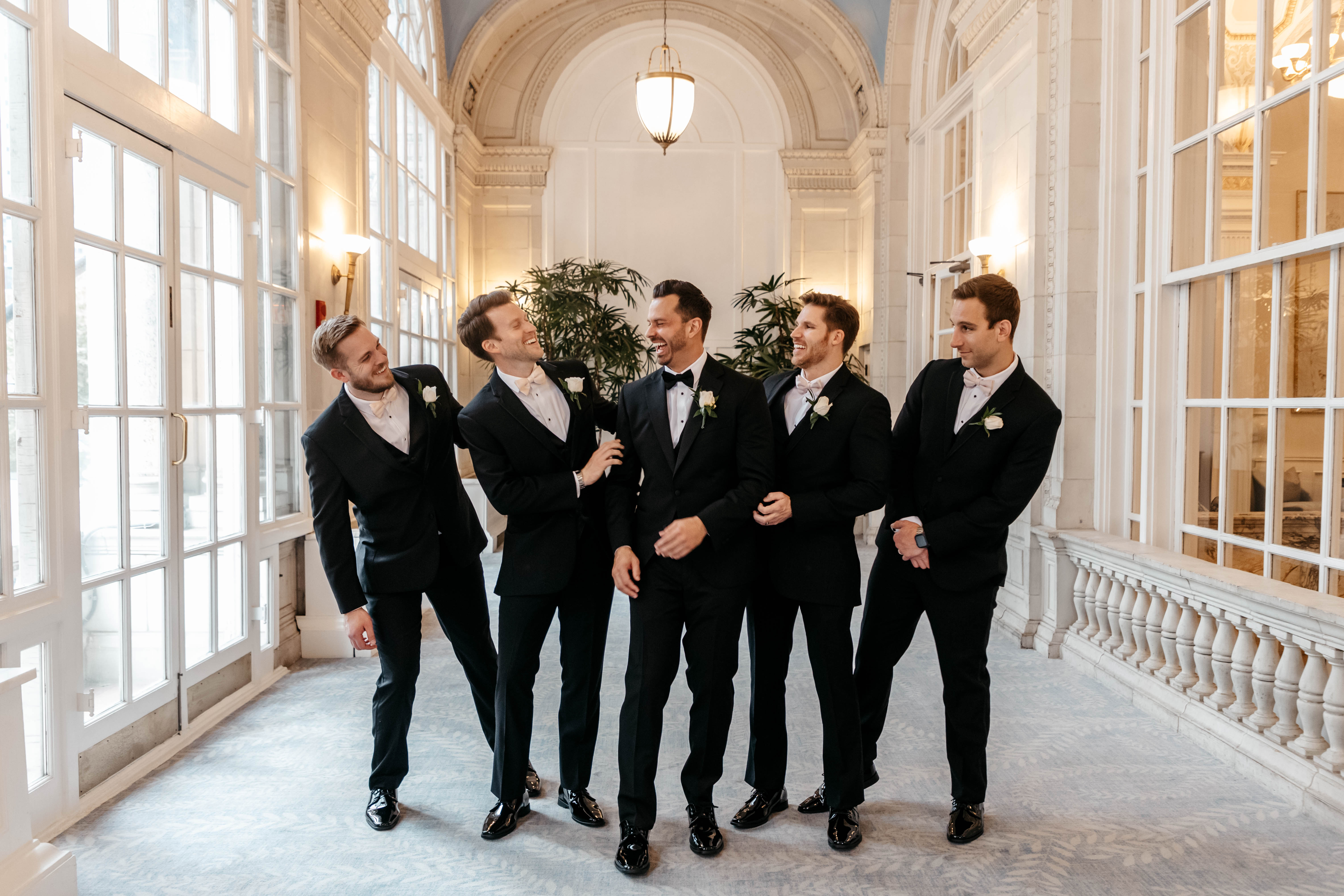 Groom and groomsmen laughing together in sunlit grand hallway with arched windows and chandelier