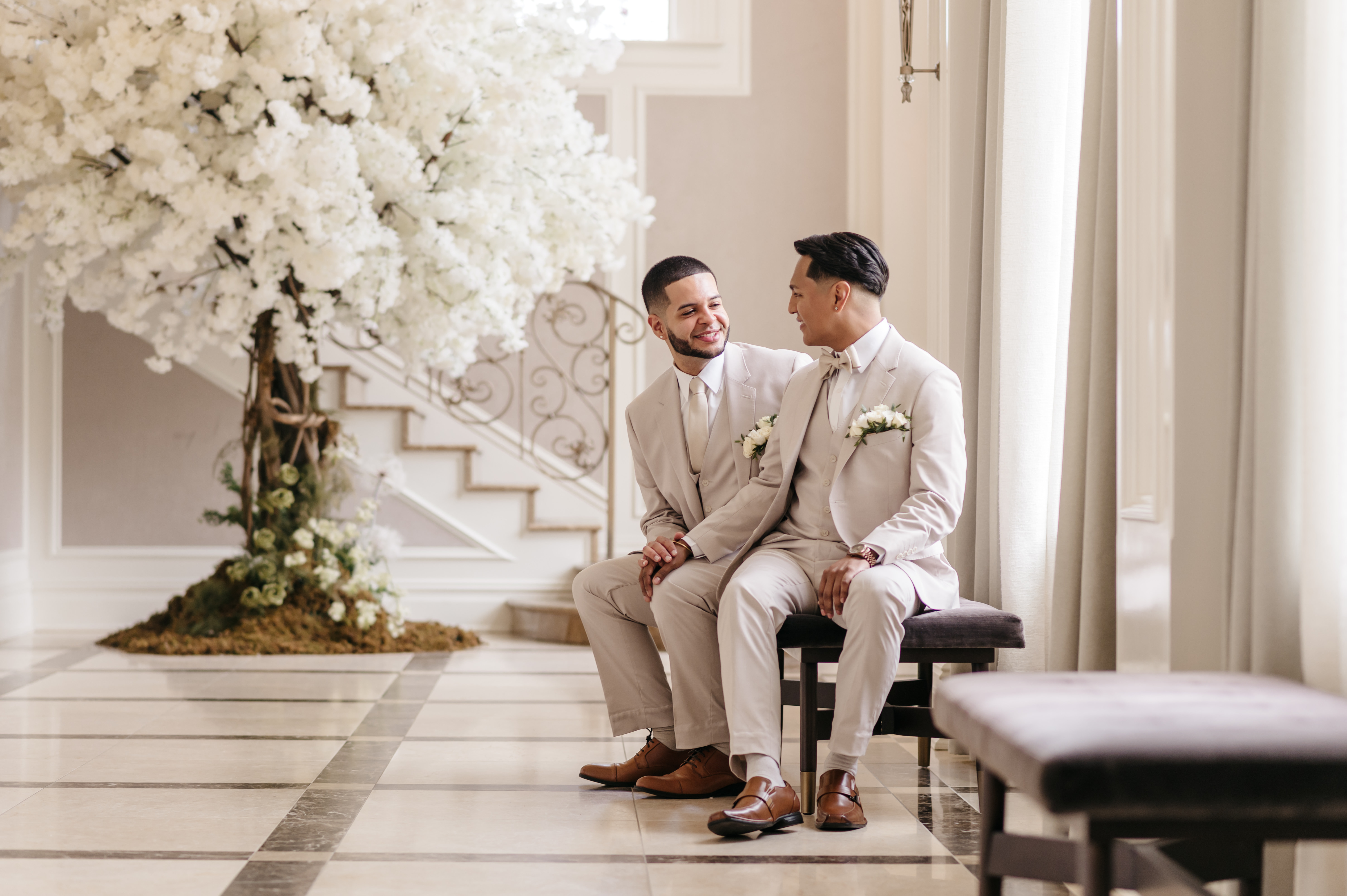 Two grooms in elegant tan suits seated together smiling under a white blossom tree