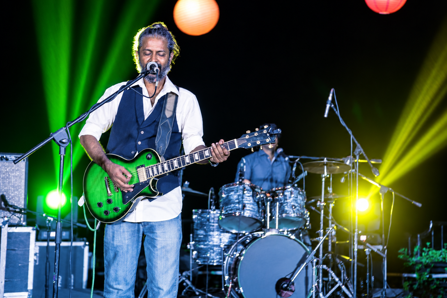 The Blue Mister performing on stage with a green guitar alongside a drummer under stage lanterns
