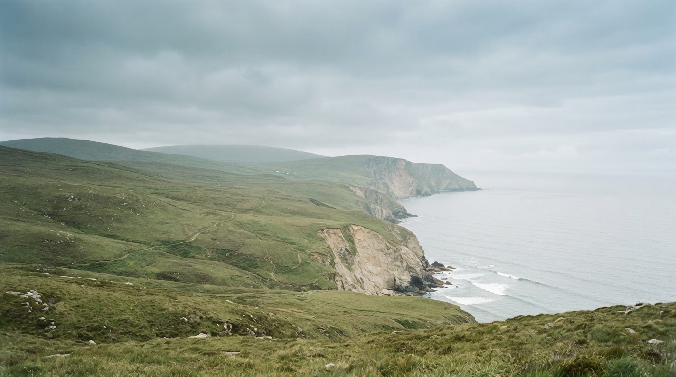 Soft green rolling hills meeting pale sea cliffs on Ireland's west coast under a muted overcast sky