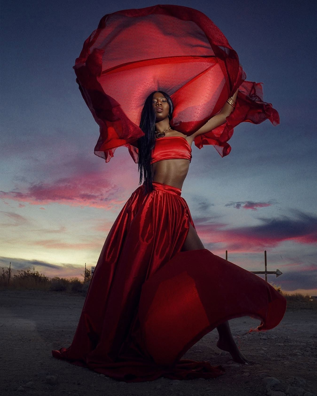 Dramatic high-fashion editorial of a woman in a billowing red satin dress against a vibrant twilight sunset