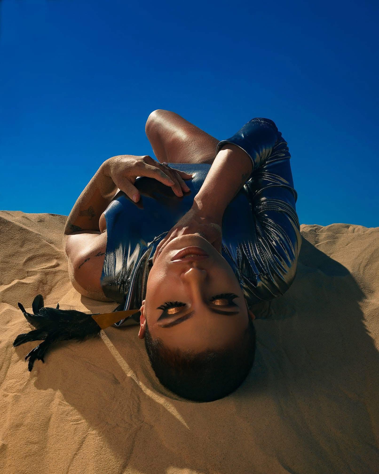Editorial portrait lying in golden dunes with metallic styling under blue sky