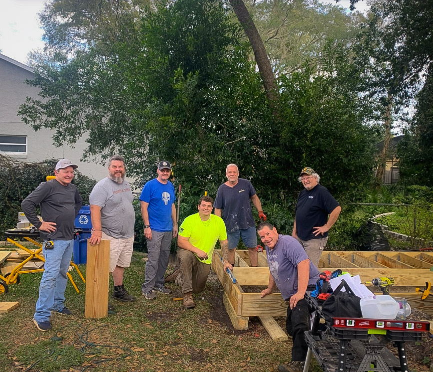 The build crew of men smiling together standing around the freshly framed deck under the tree