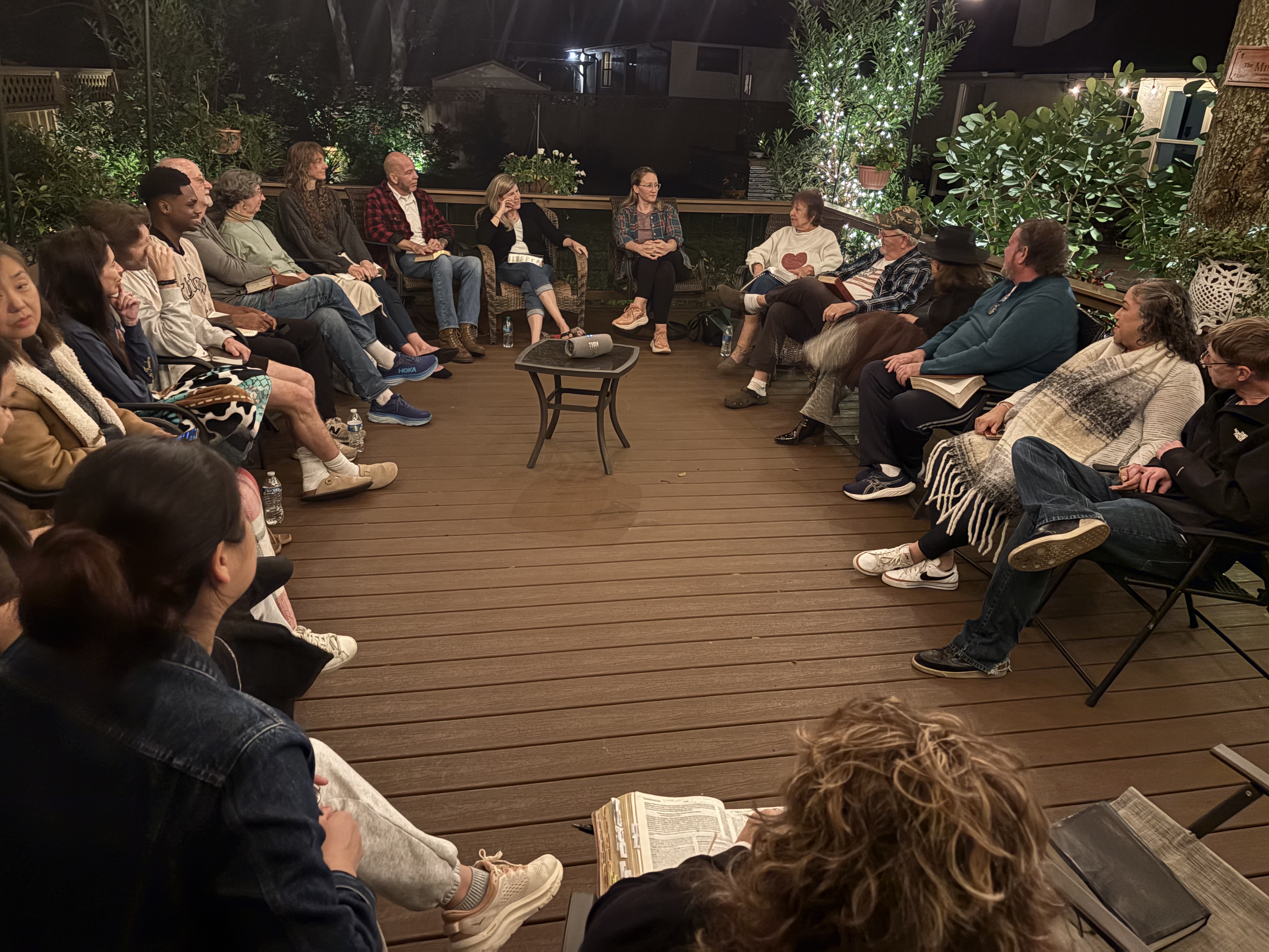 Believers sitting in a circle on the deck with open Bibles under the trees and string lights
