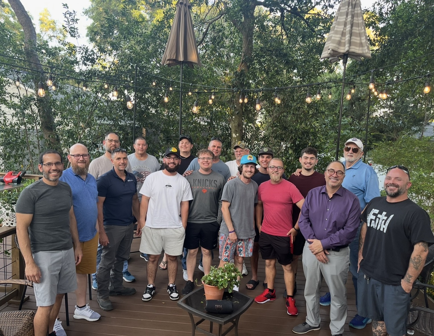 Men gathered together on the deck under the glowing string lights and shade tree branches