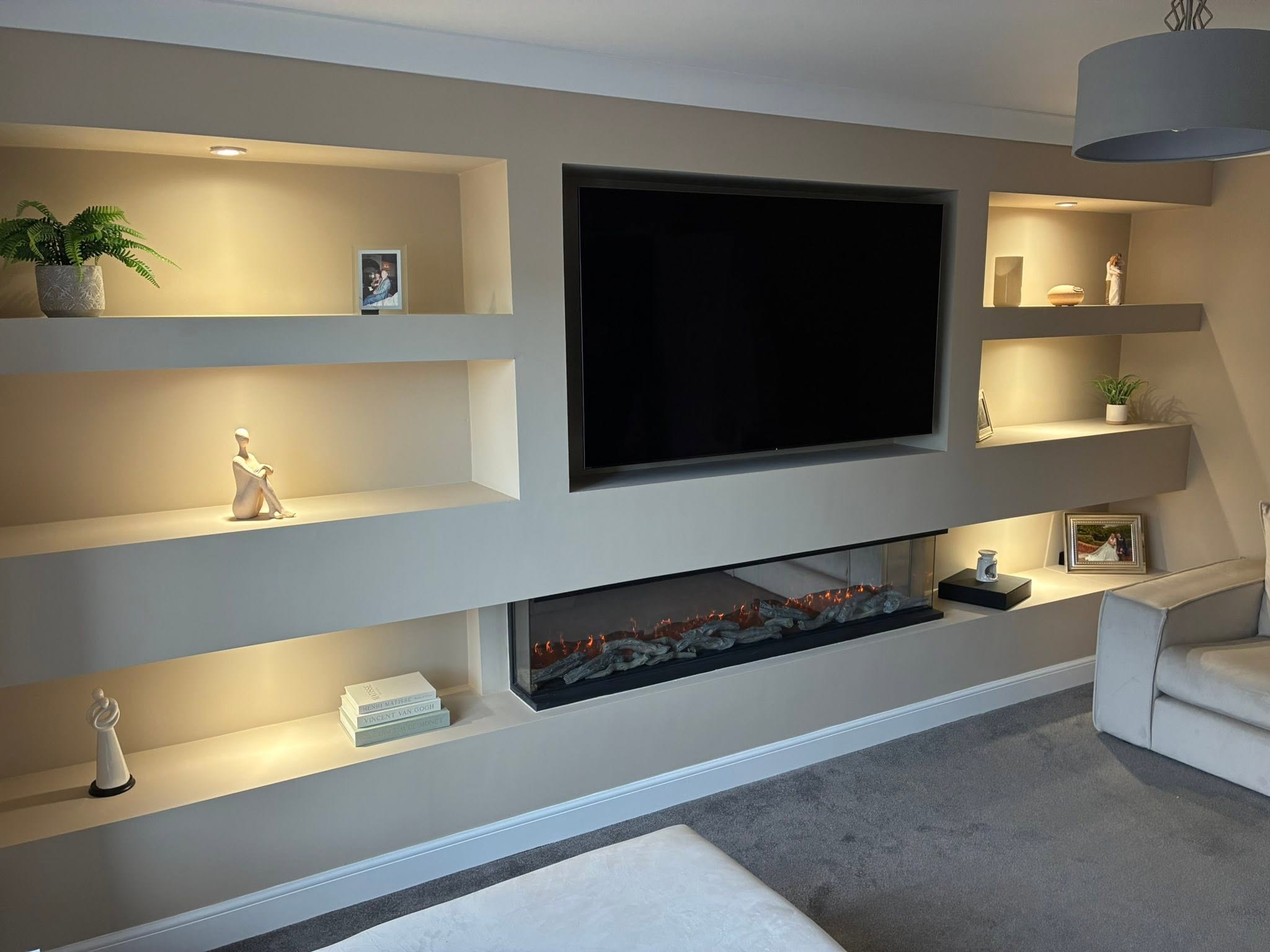 Full-width bespoke media wall with warm recessed LED alcove lighting, flush TV recess and panoramic electric flame fireplace by SJ Joinery & Building