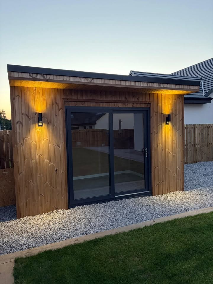 Bespoke timber garden studio with warm exterior sconce lighting at dusk by SJ Joinery & Building