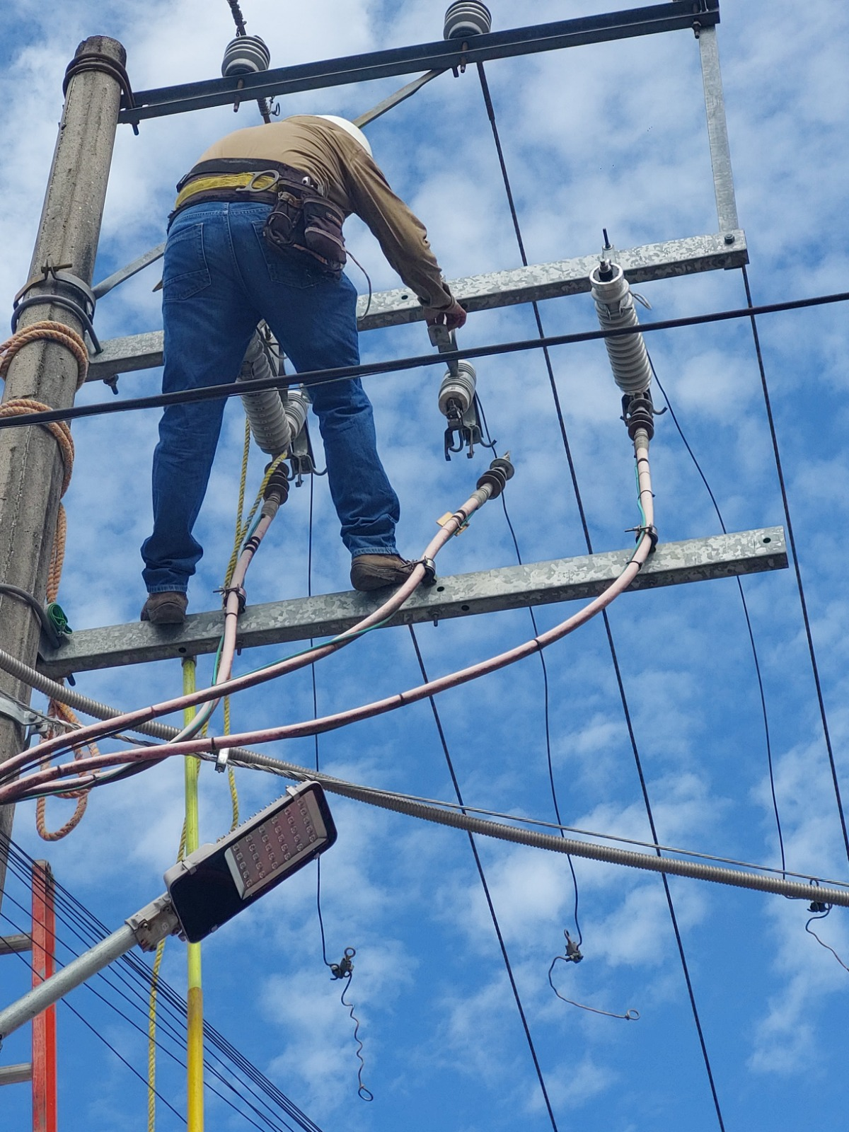 Liniero electricista en altura trabajando sobre cruceta con cortacircuitos y aisladores en poste de distribución
