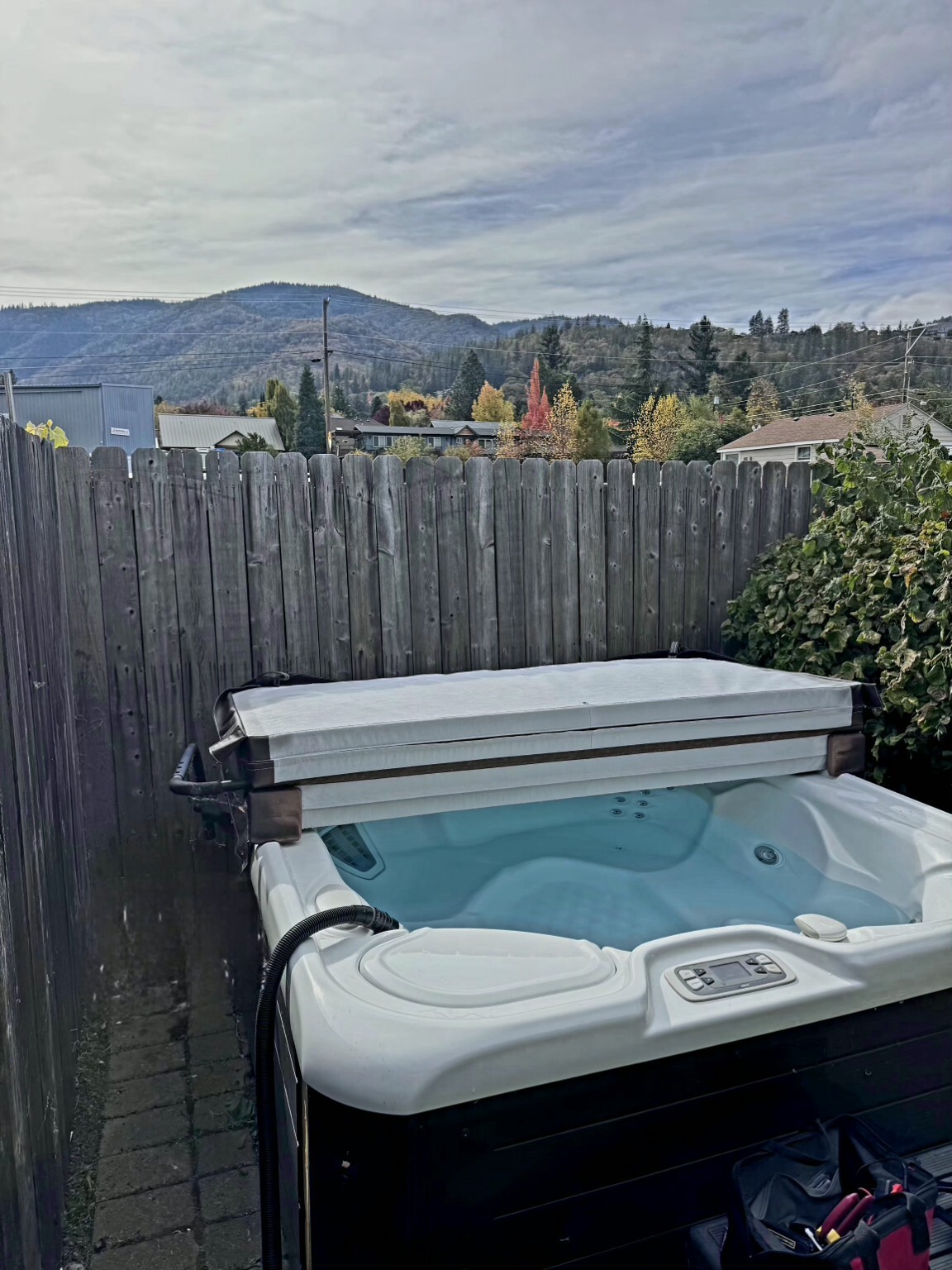 Outdoor hot tub with folded cover against a wooden privacy fence with hillside autumn trees in the background